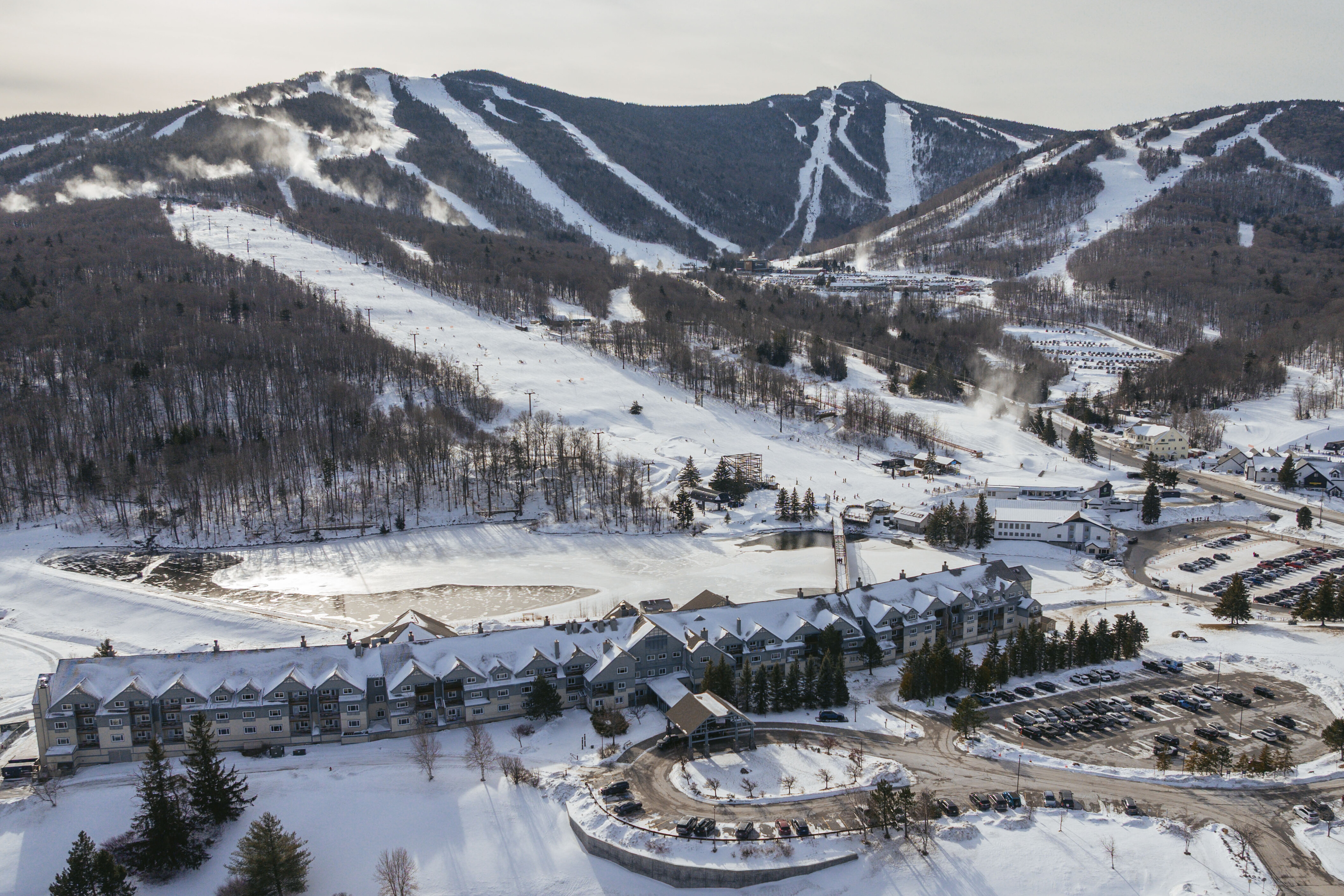 Aerial view of a snowy ski resort with mountains, a lake, and parking areas.