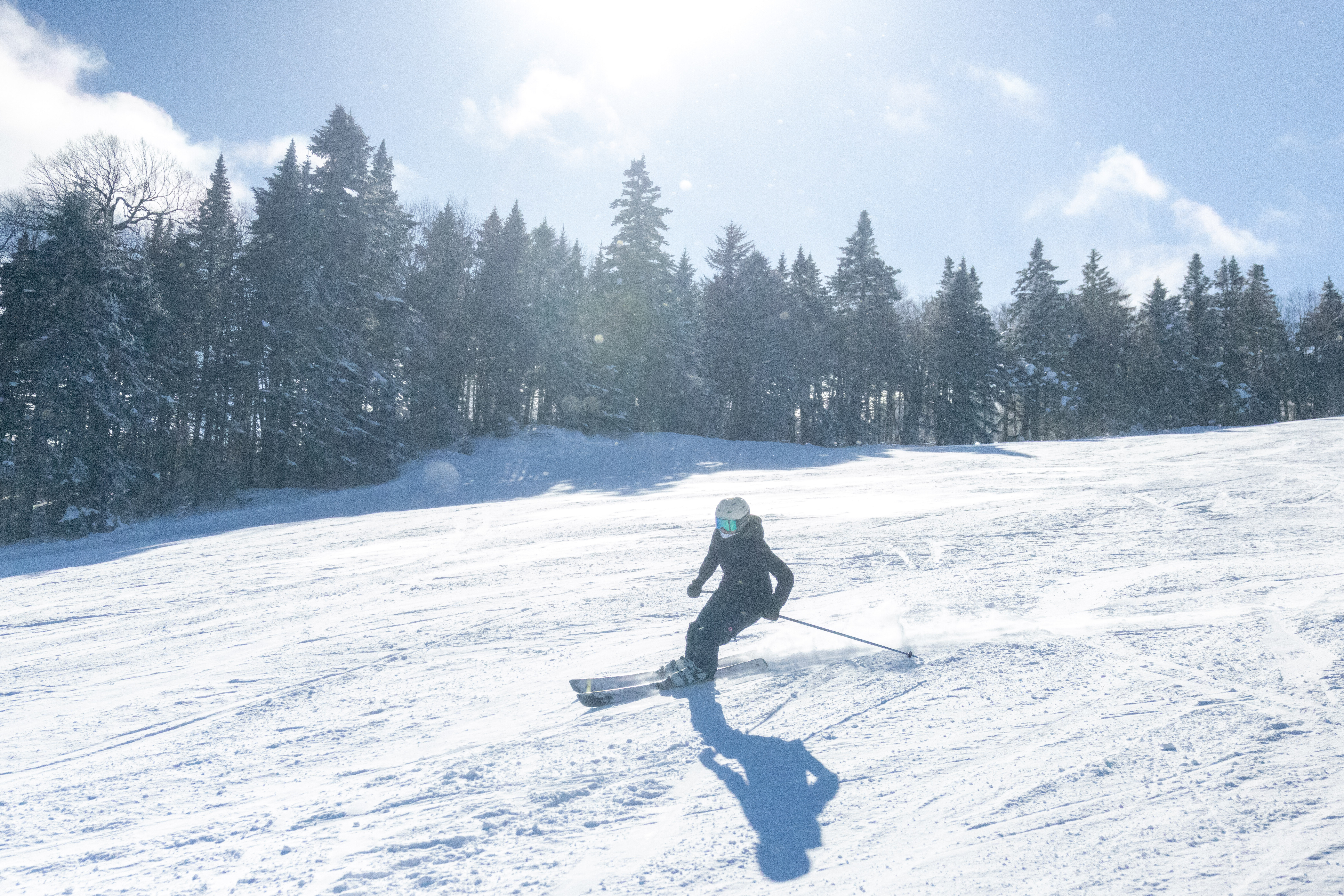 A skier glides down a snowy slope with trees and sun in the background.