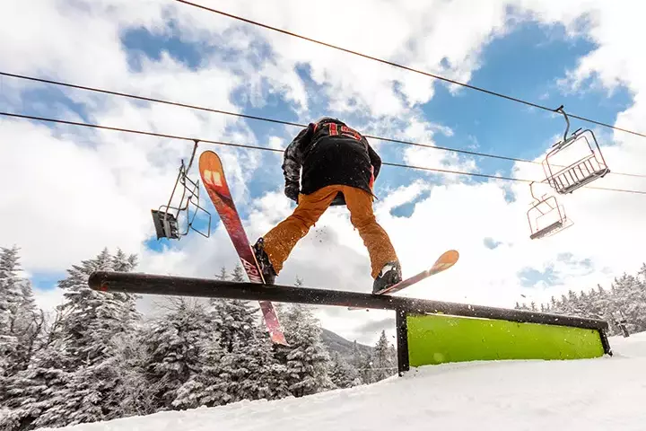 Skier performing a trick on a rail with snowy trees and a chairlift in the background.