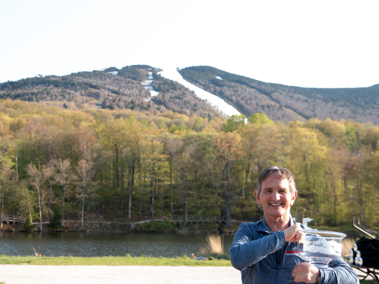 Smiling man holding a trophy with mountains and trees in the background.