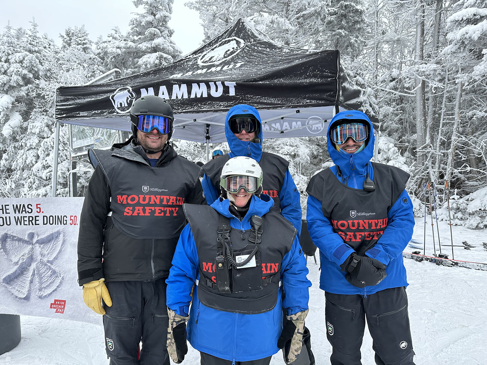 Four Mountain Safety members in winter gear stand near a tent in snowy terrain.