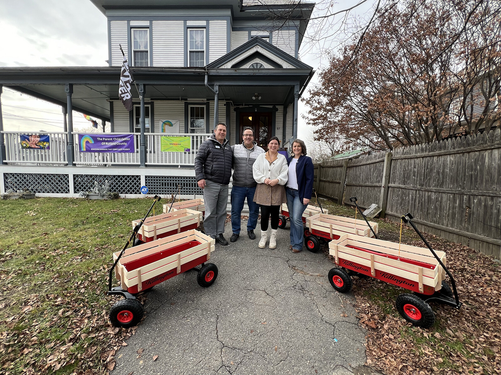 Four people stand in front of a house with red-and-white wagons in a grassy area.