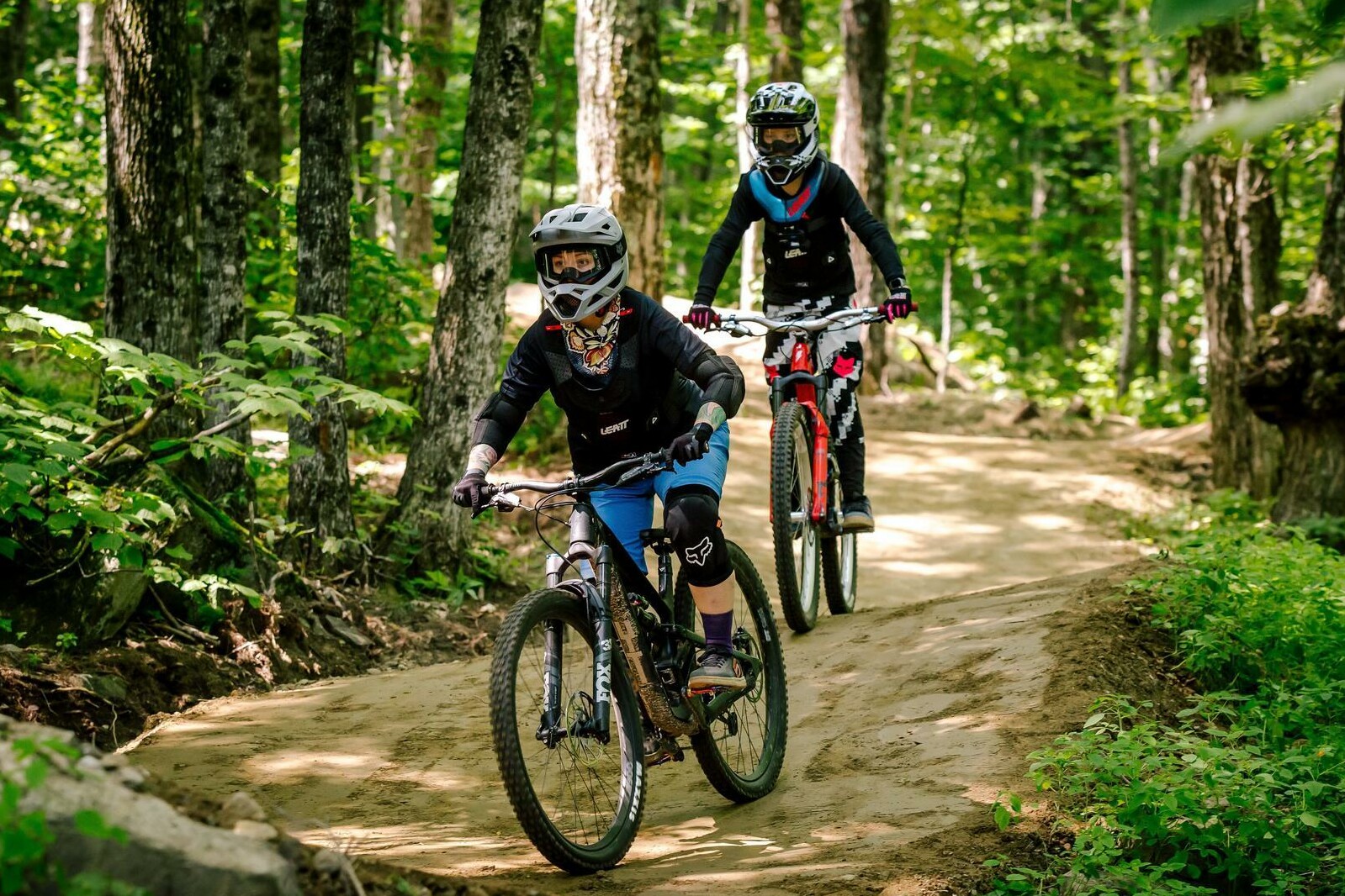 Two bikers navigate a dirt trail through a lush, green forest.