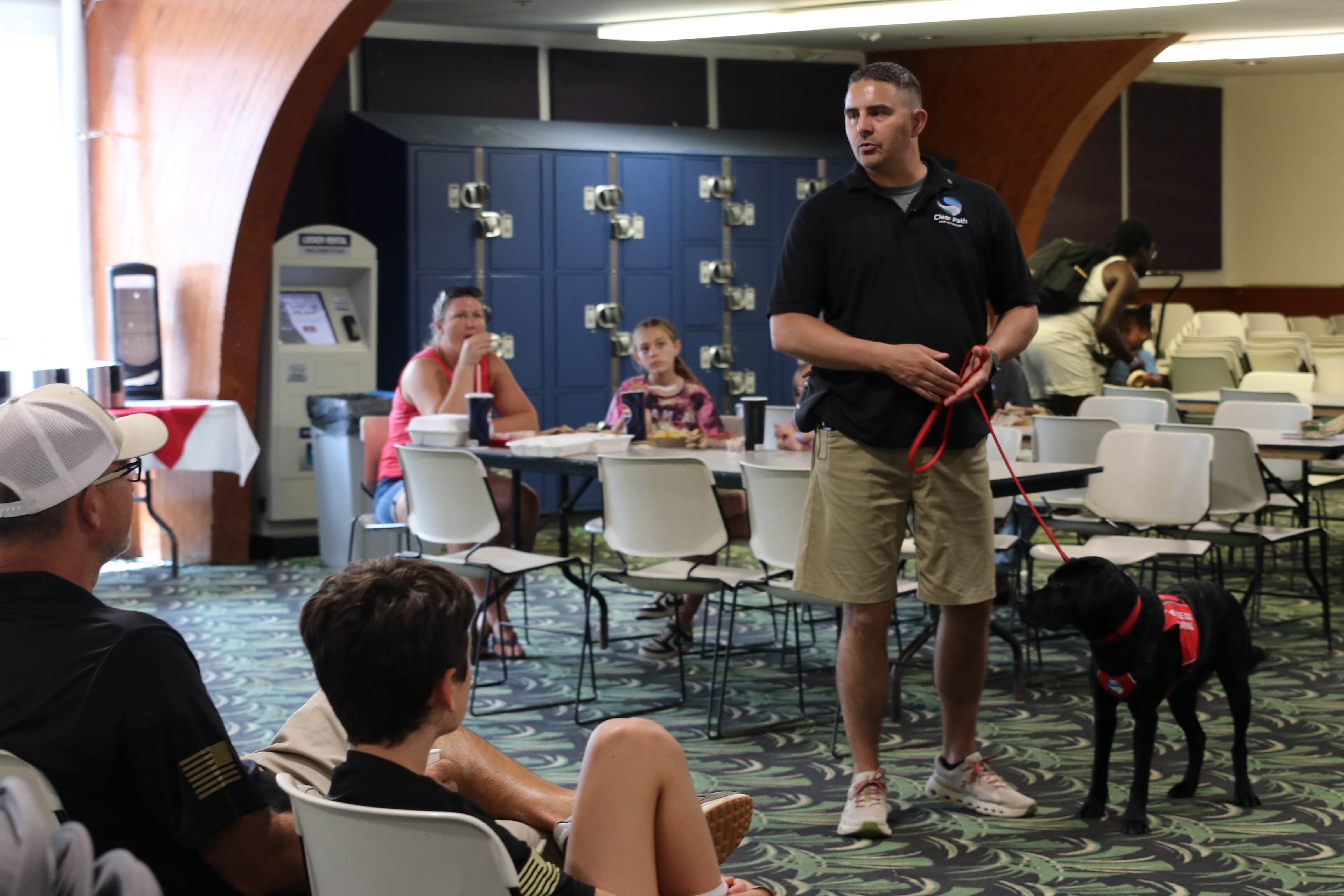 A man speaks to an audience while handling a service dog.