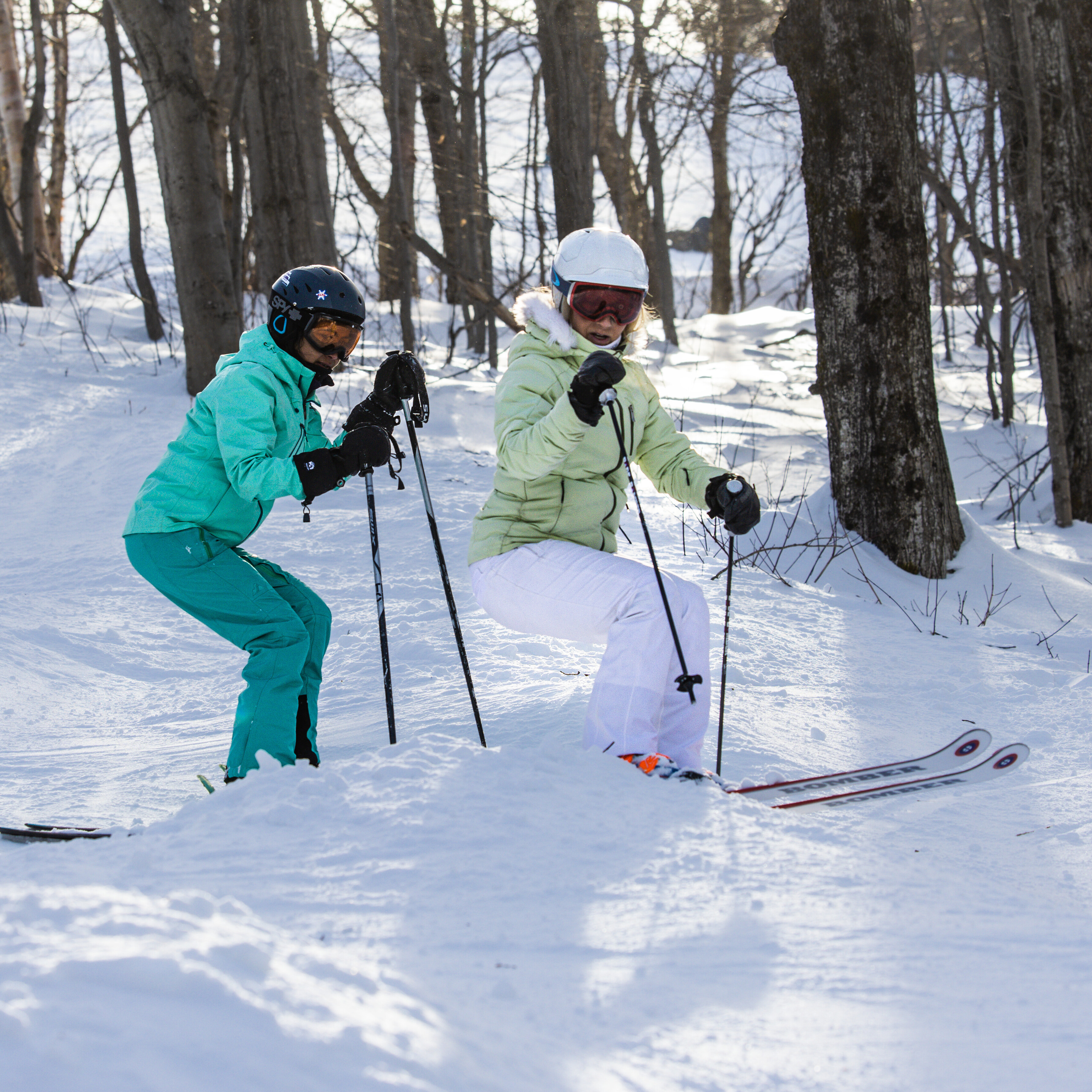 Two skiers navigating snowy terrain among trees, enjoying a sunny day on the slopes.