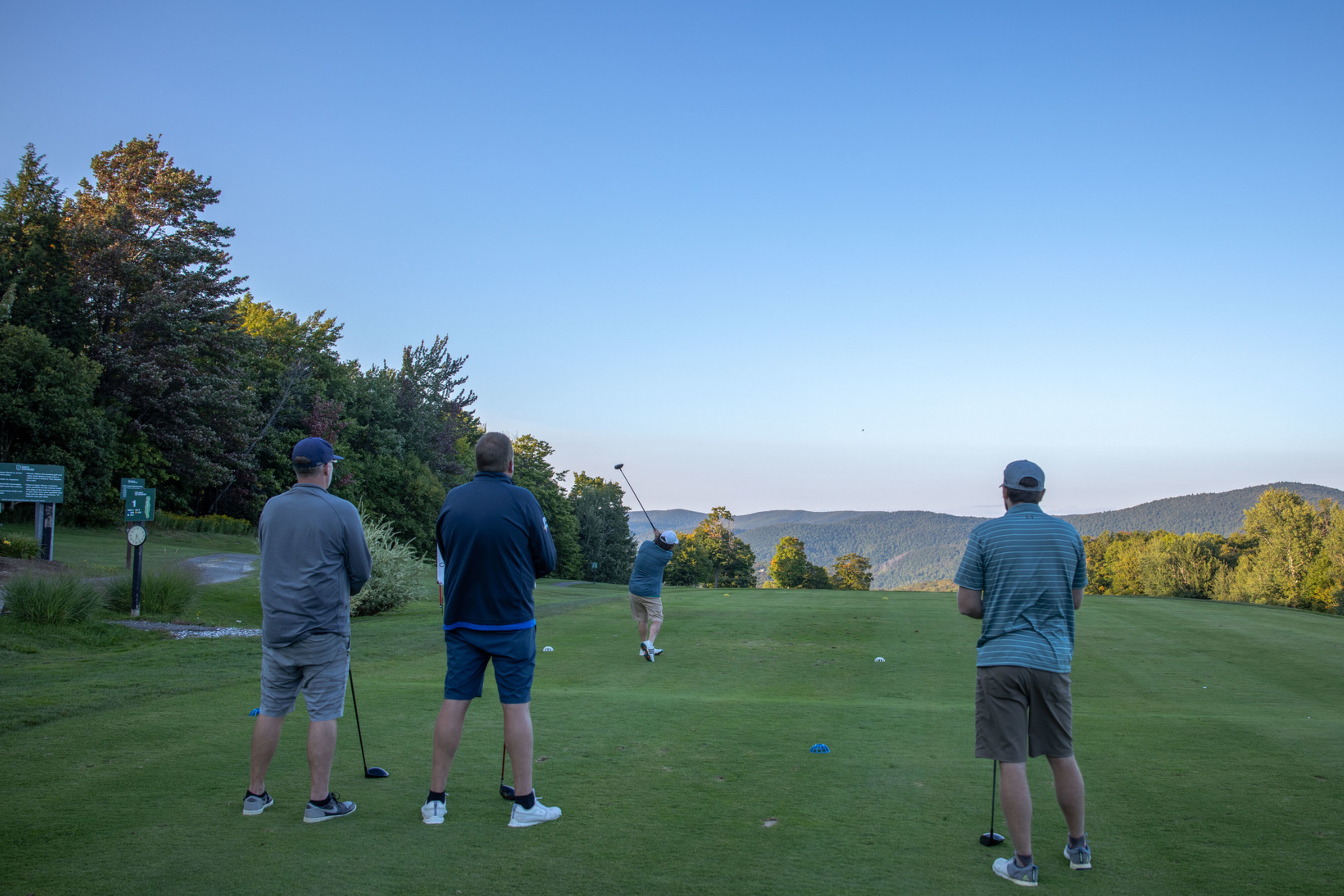 Four golfers on a green tee, watching one player drive a golf ball toward mountains.