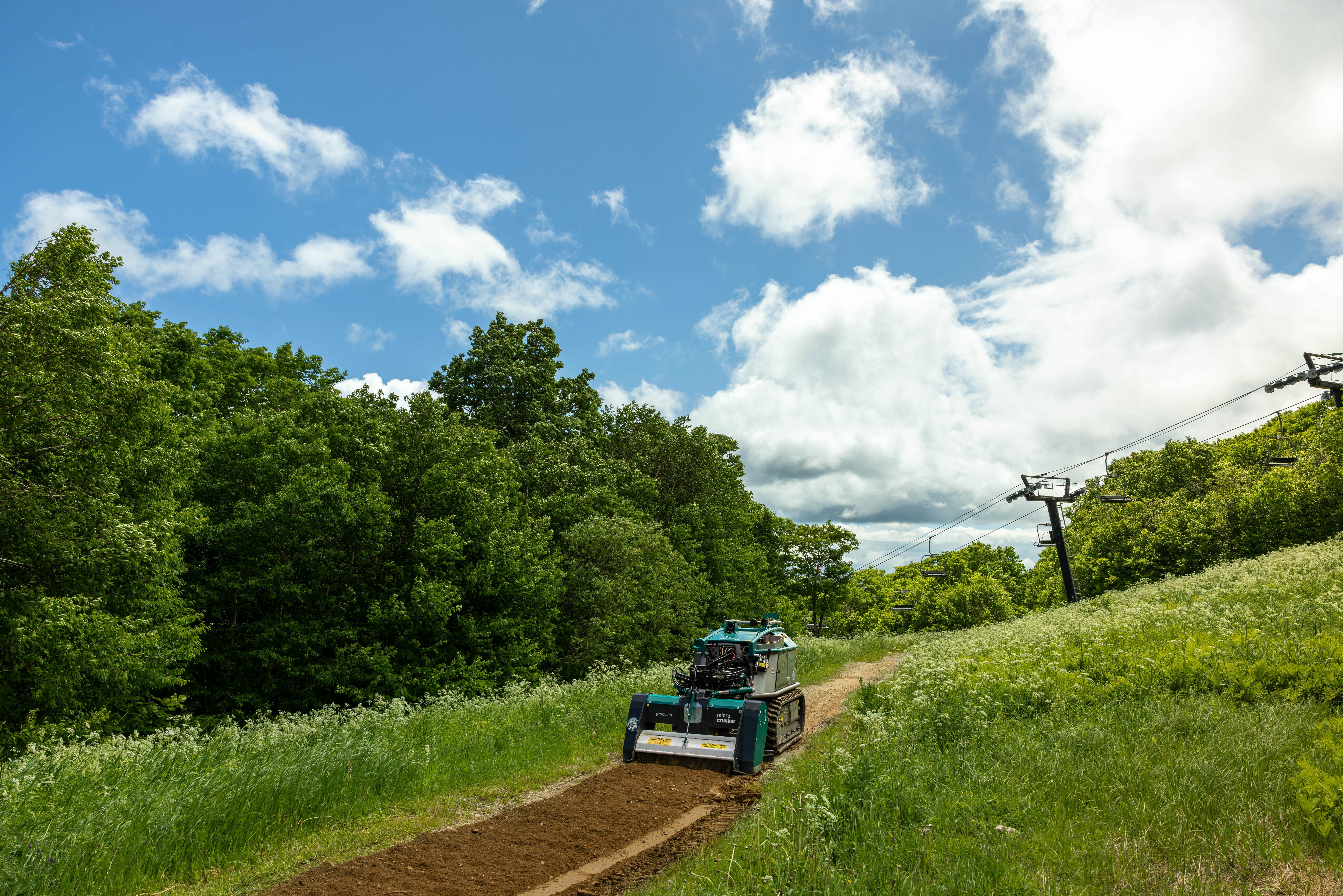 A machine leveling a mountain bike trail surrounded by green trees and grass under a blue sky.