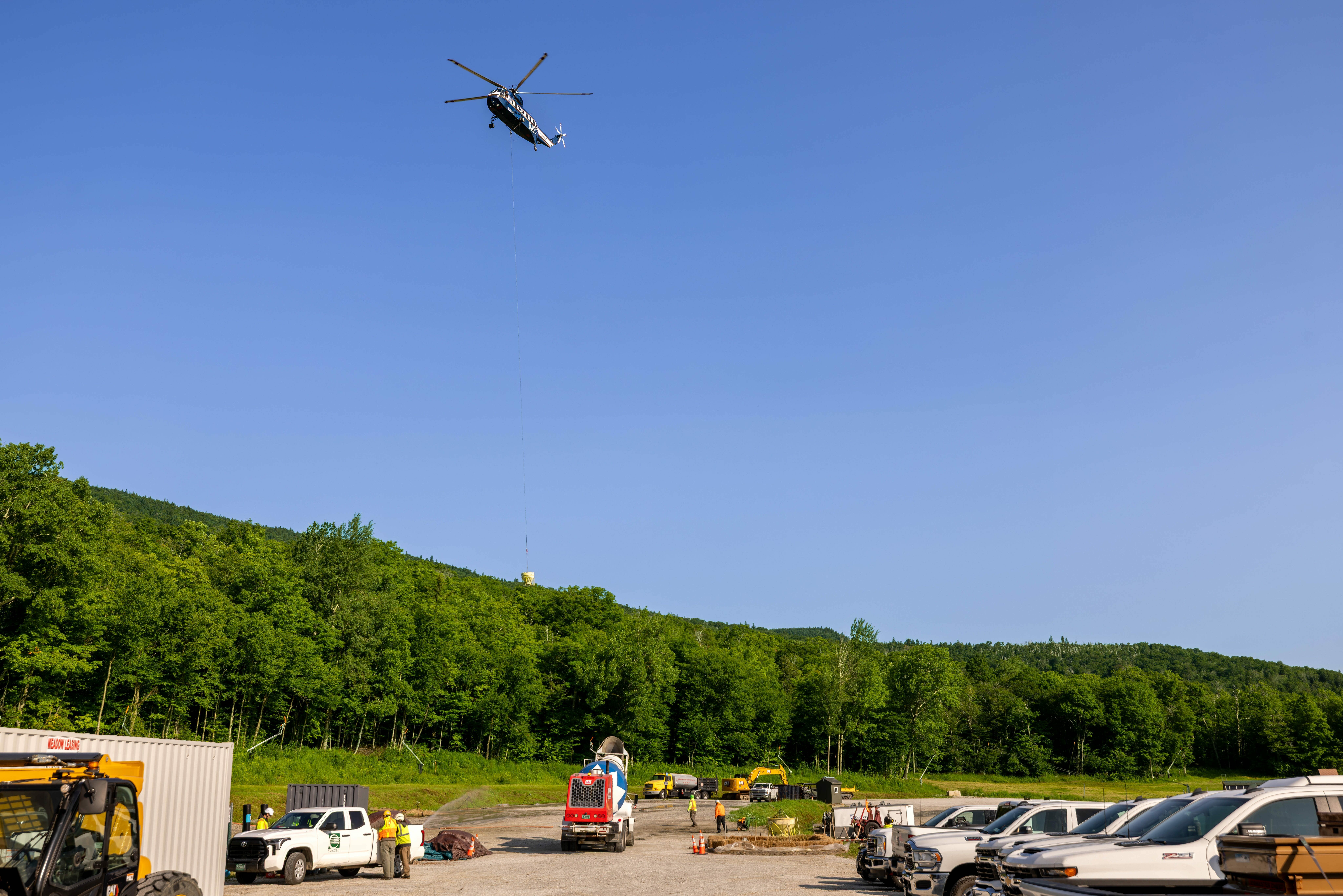 Helicopter flying overhead, lowering a load near vehicles and greenery.