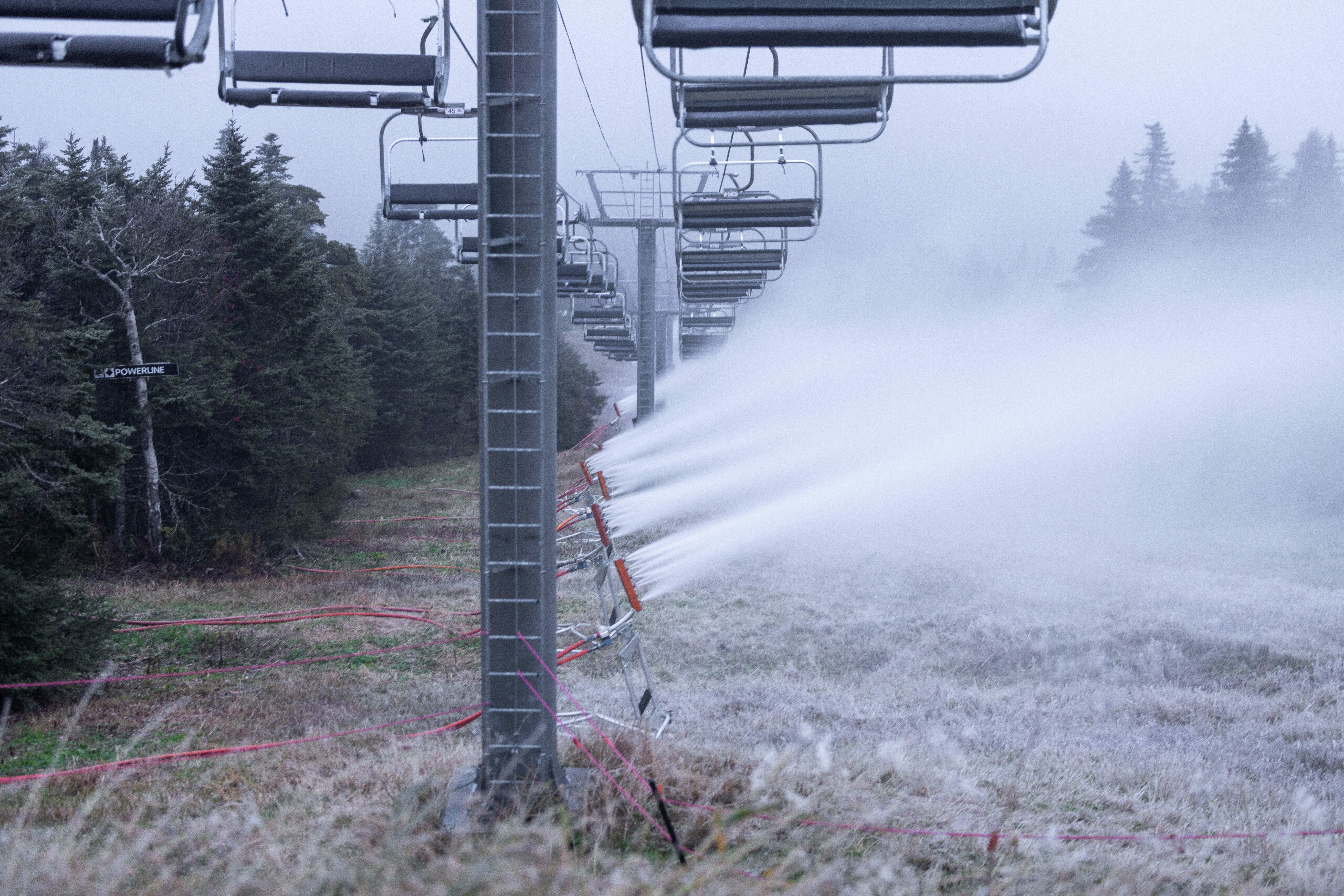 Snowmaking machines operate along a ski lift amidst foggy conditions.