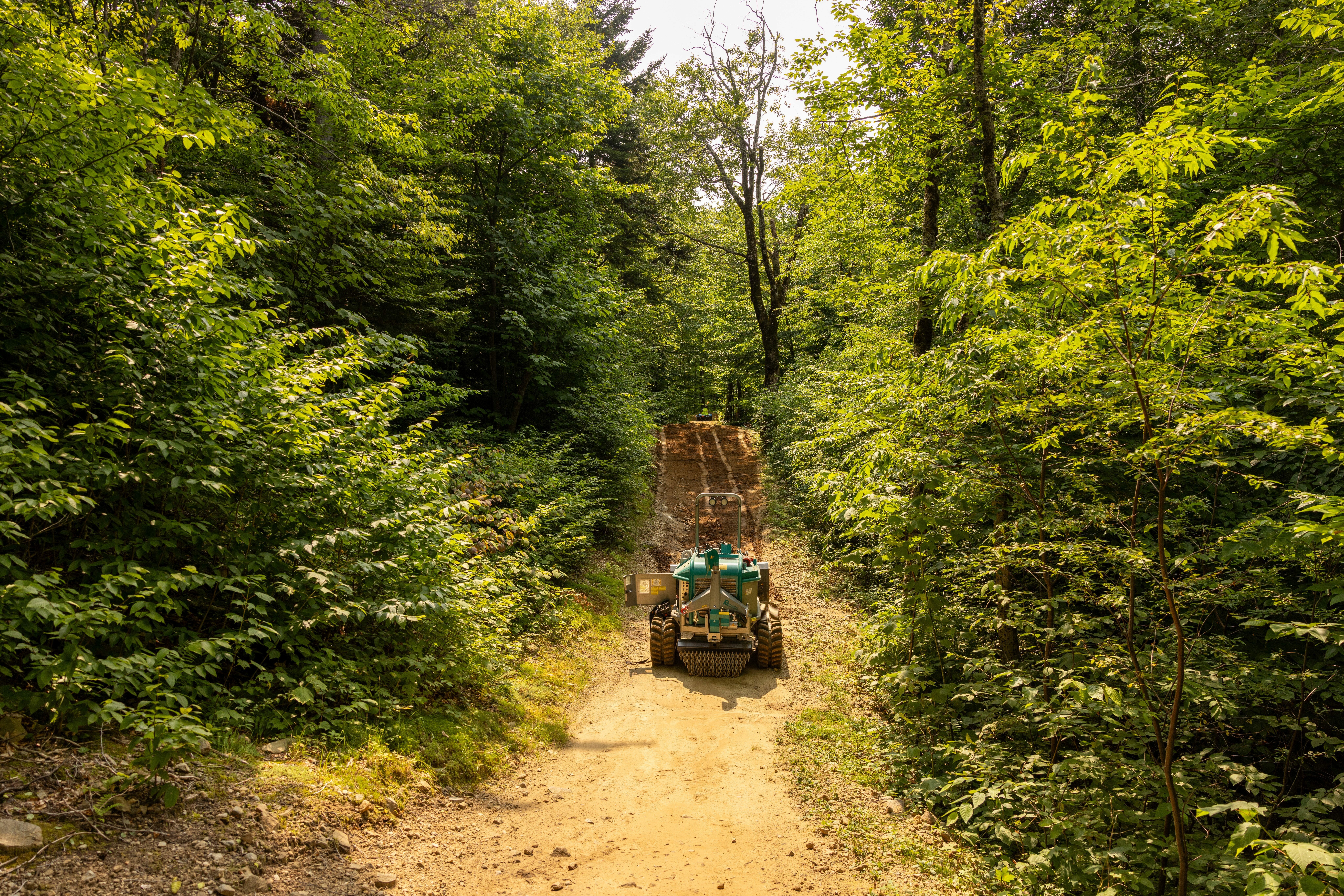 A green machine working on a mountain bike trail surrounded by lush trees.