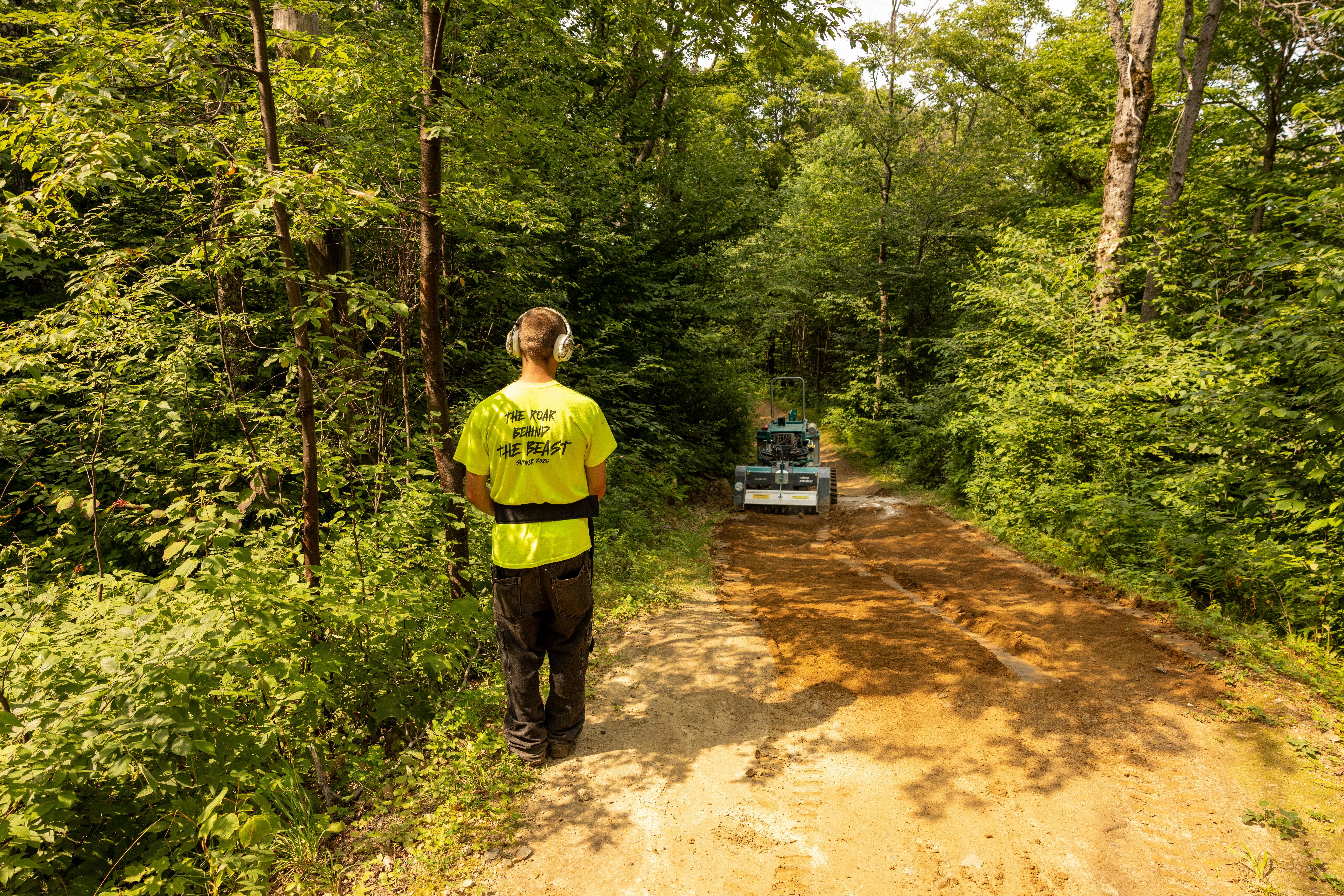 A man in a bright shirt oversees a machine on a mountain bike trail.
