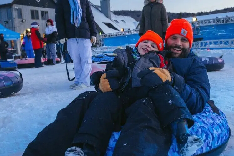 A smiling man and child sit together on a snow tube, both wearing bright orange hats.