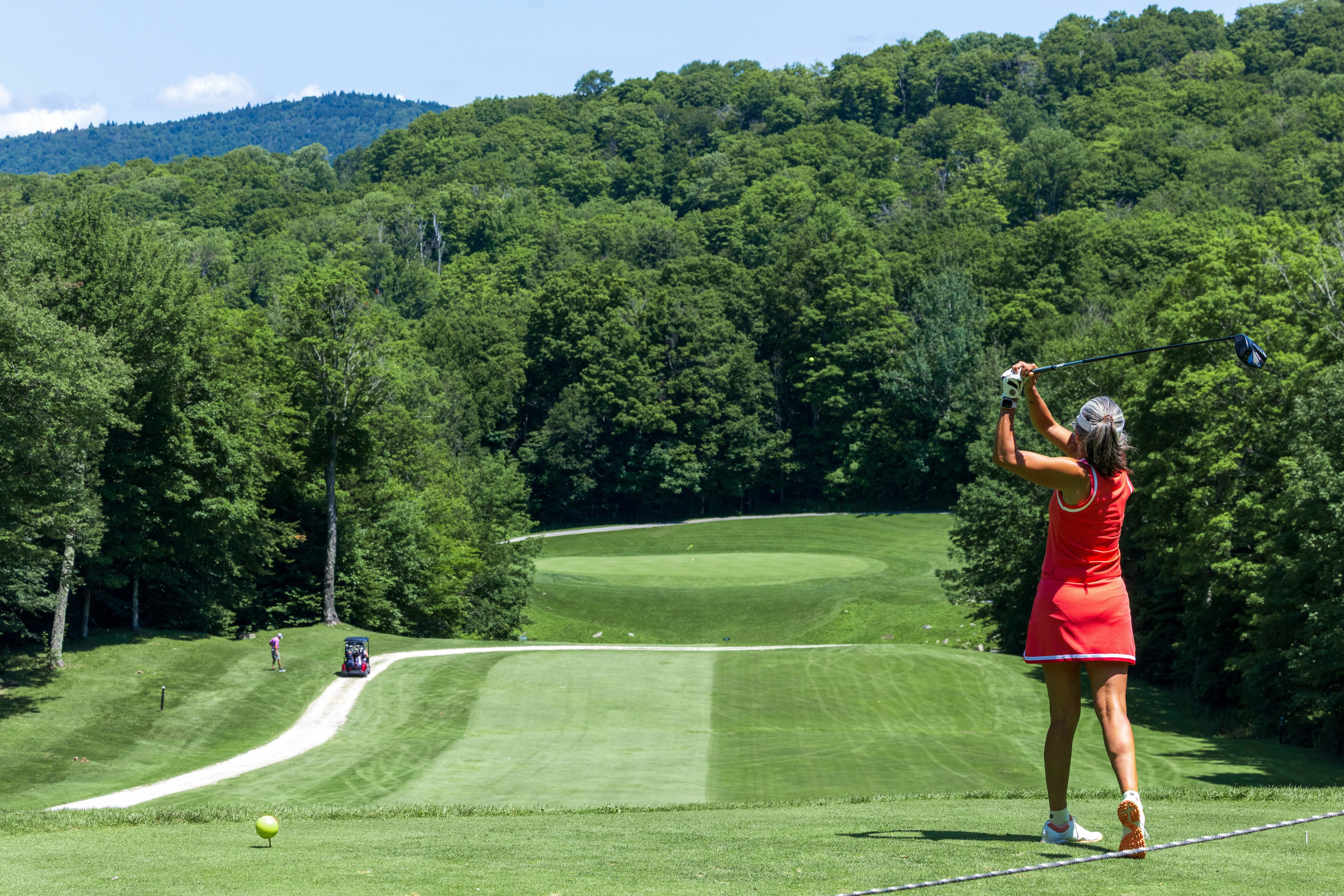 Golfer in red swings on a sunny day, green landscape and trees in the background.