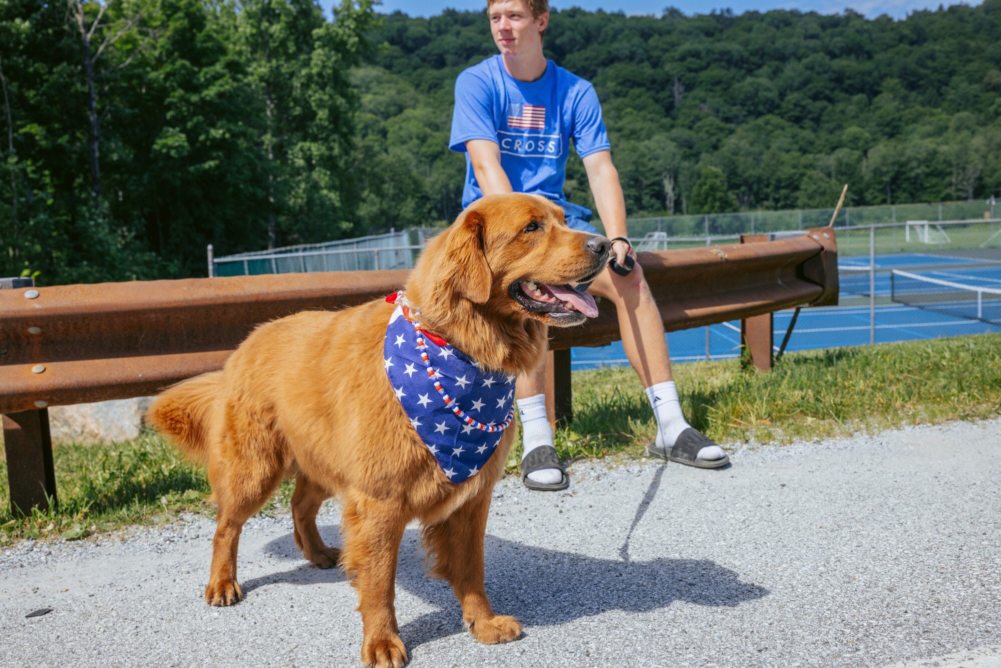 A golden retriever wearing an American flag bandana stands beside a young man outdoors.