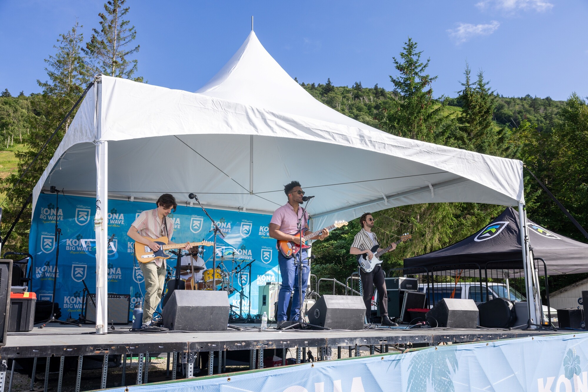 A band performs on stage under a canopy at an outdoor event surrounded by trees.