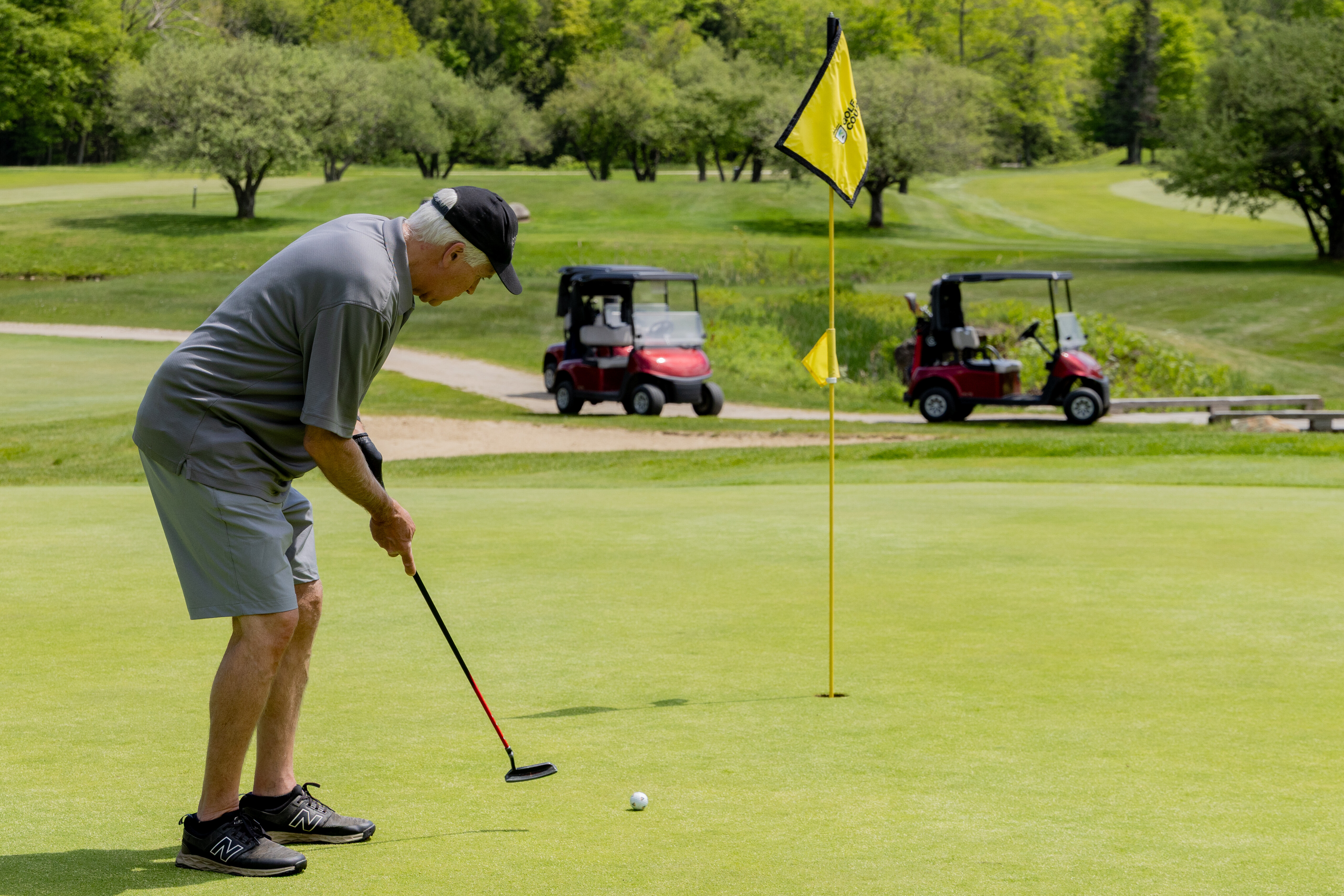 An elderly man putts on a golf green with carts in the background.