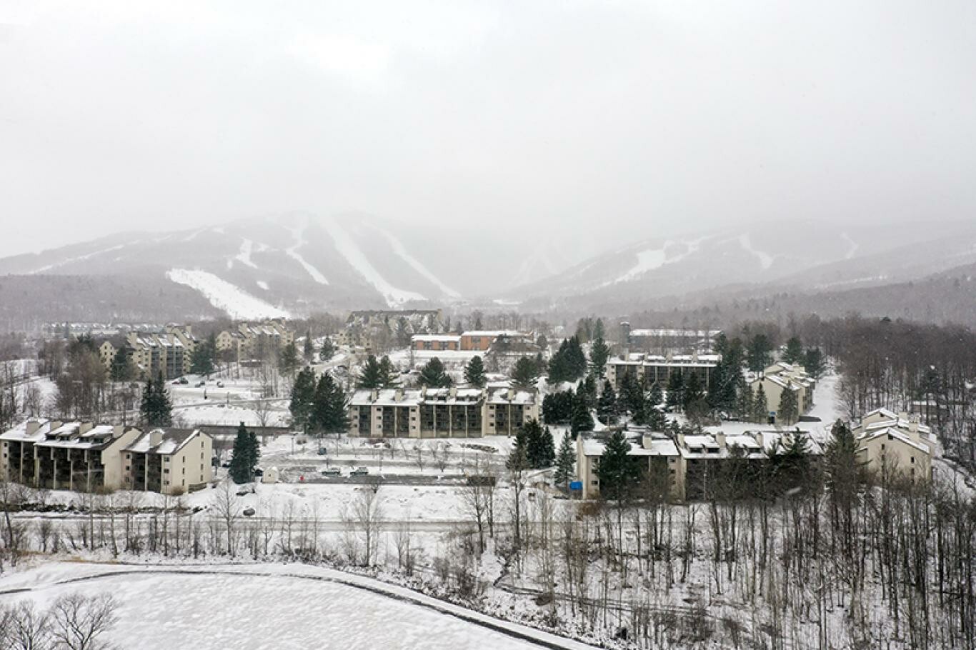 Snow-covered mountain resort with buildings and trees, under a cloudy sky.