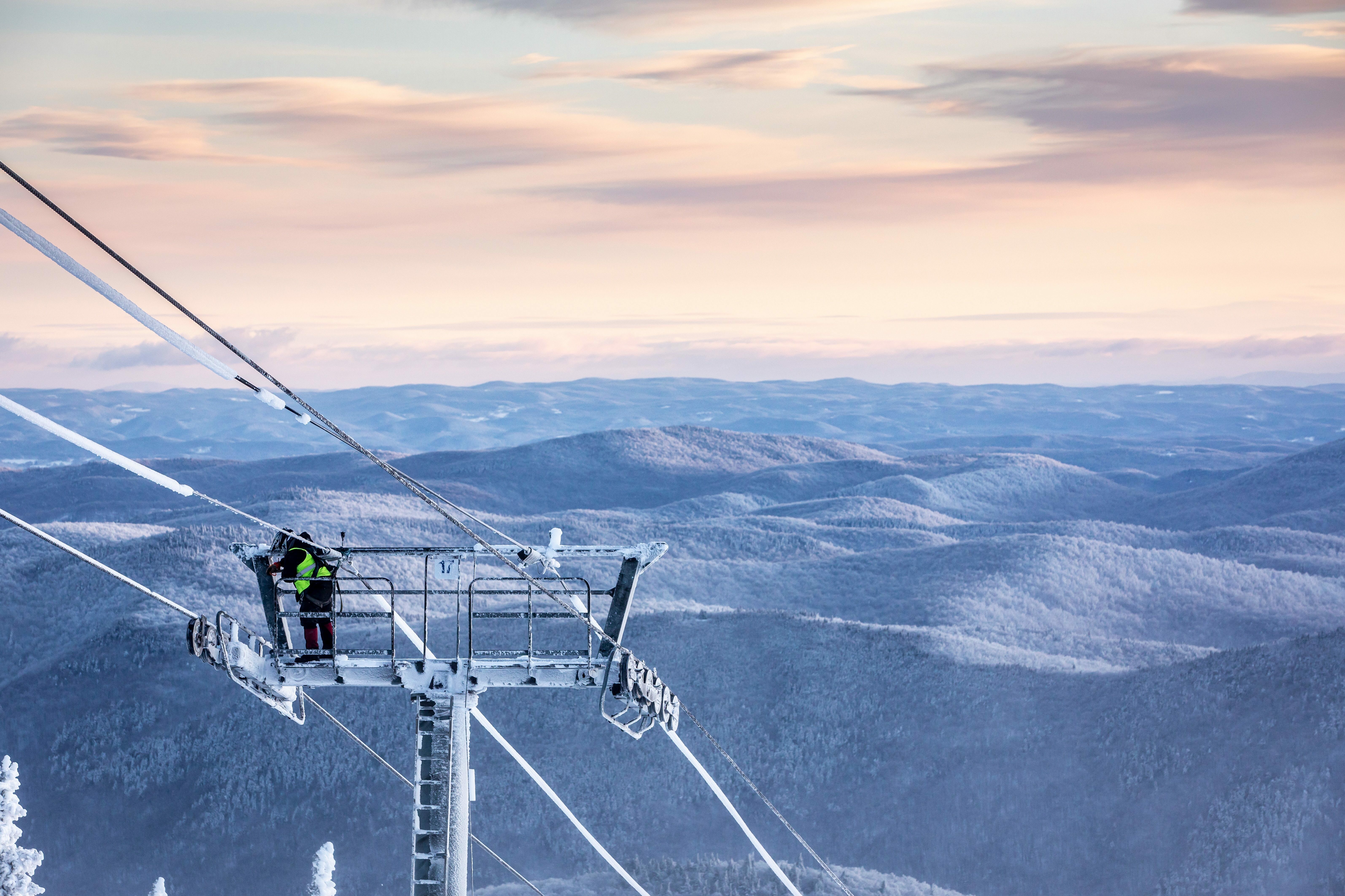 Worker on a snowy ski lift tower with mountainous landscape in the background.