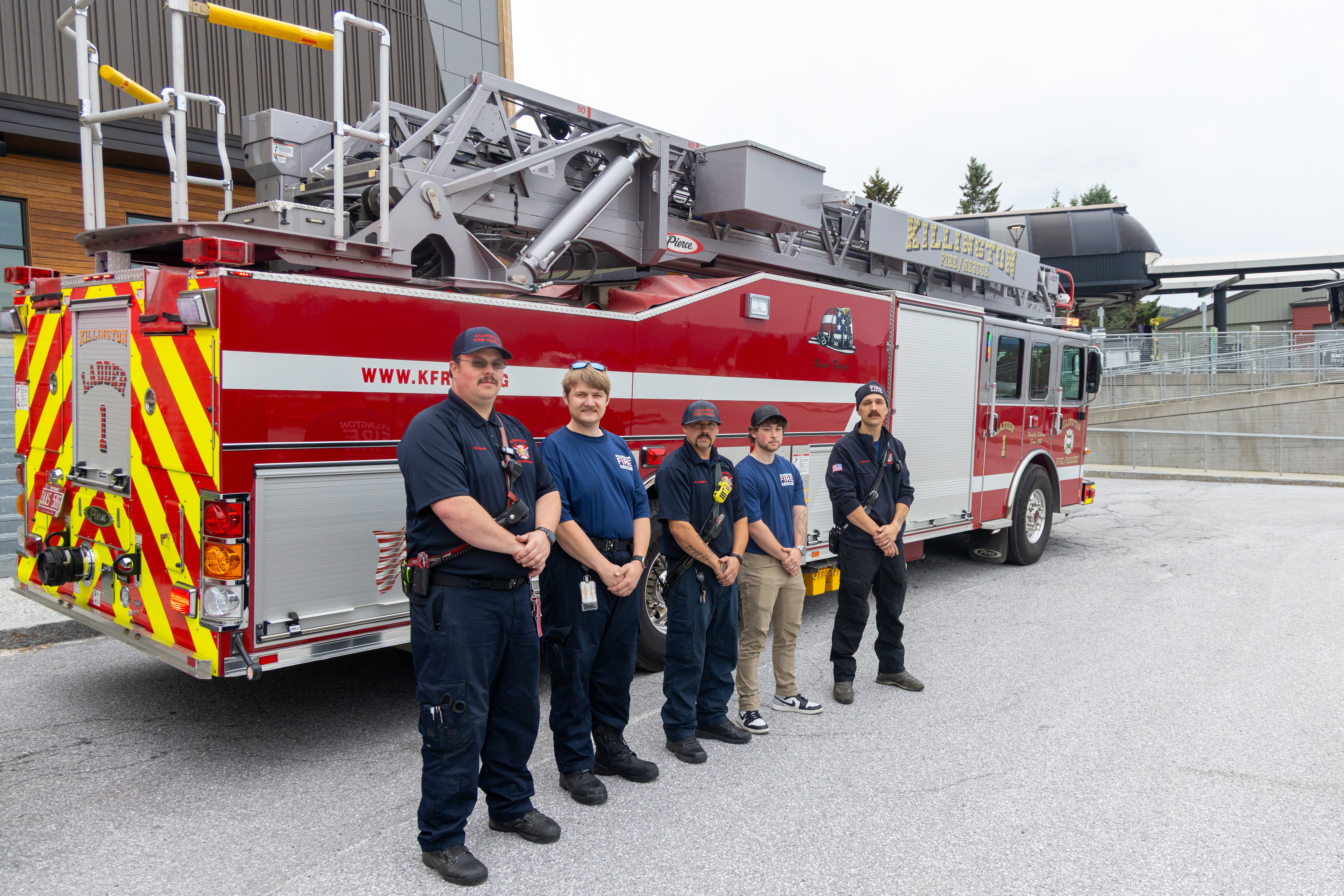 Five firefighters pose in front of a fire truck in a parking lot.