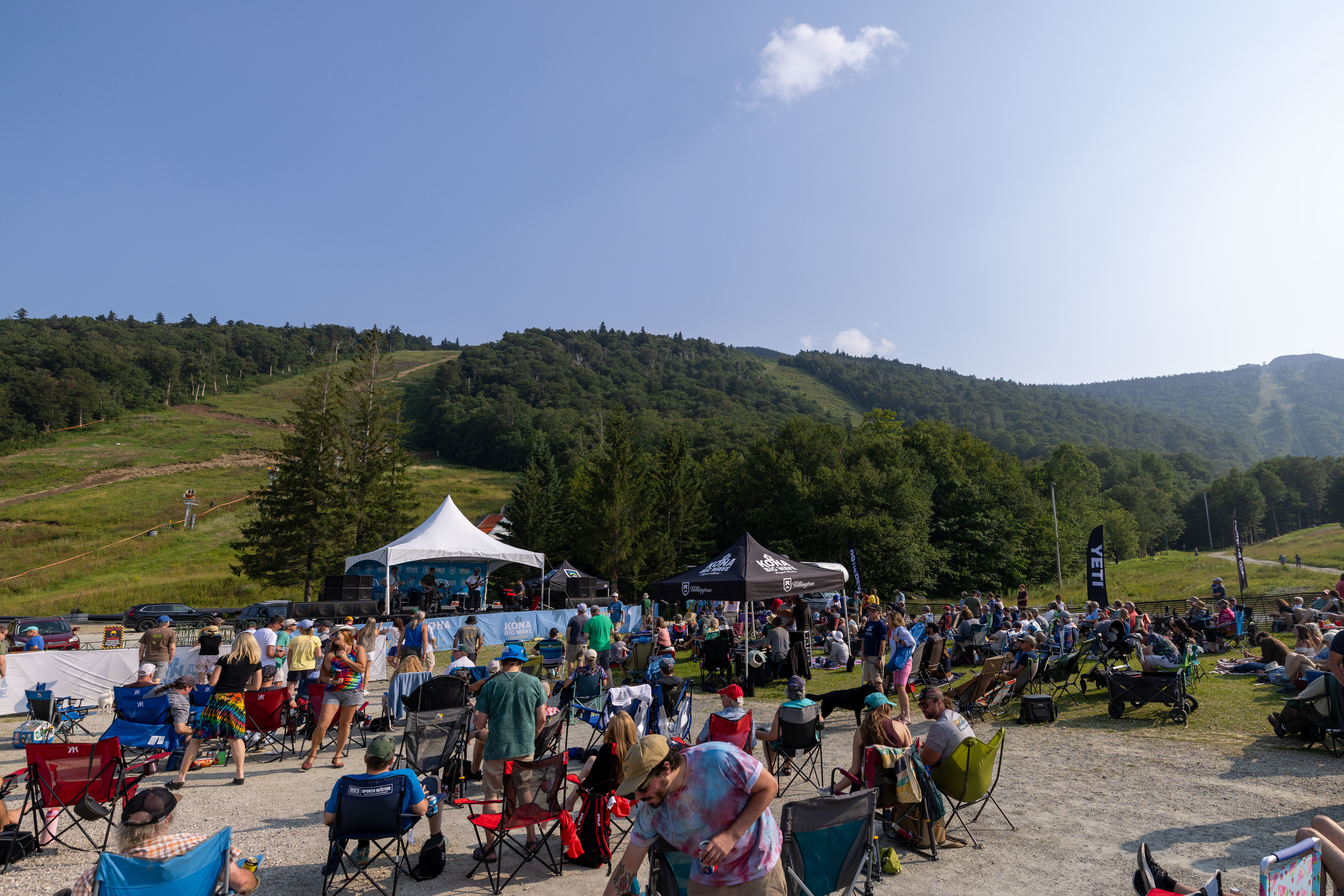 Outdoor music festival with a crowd seated on the grass, near a mountain backdrop.