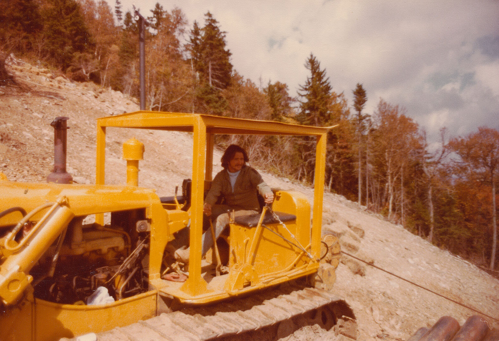 Man operating a yellow bulldozer on a hillside, surrounded by trees.