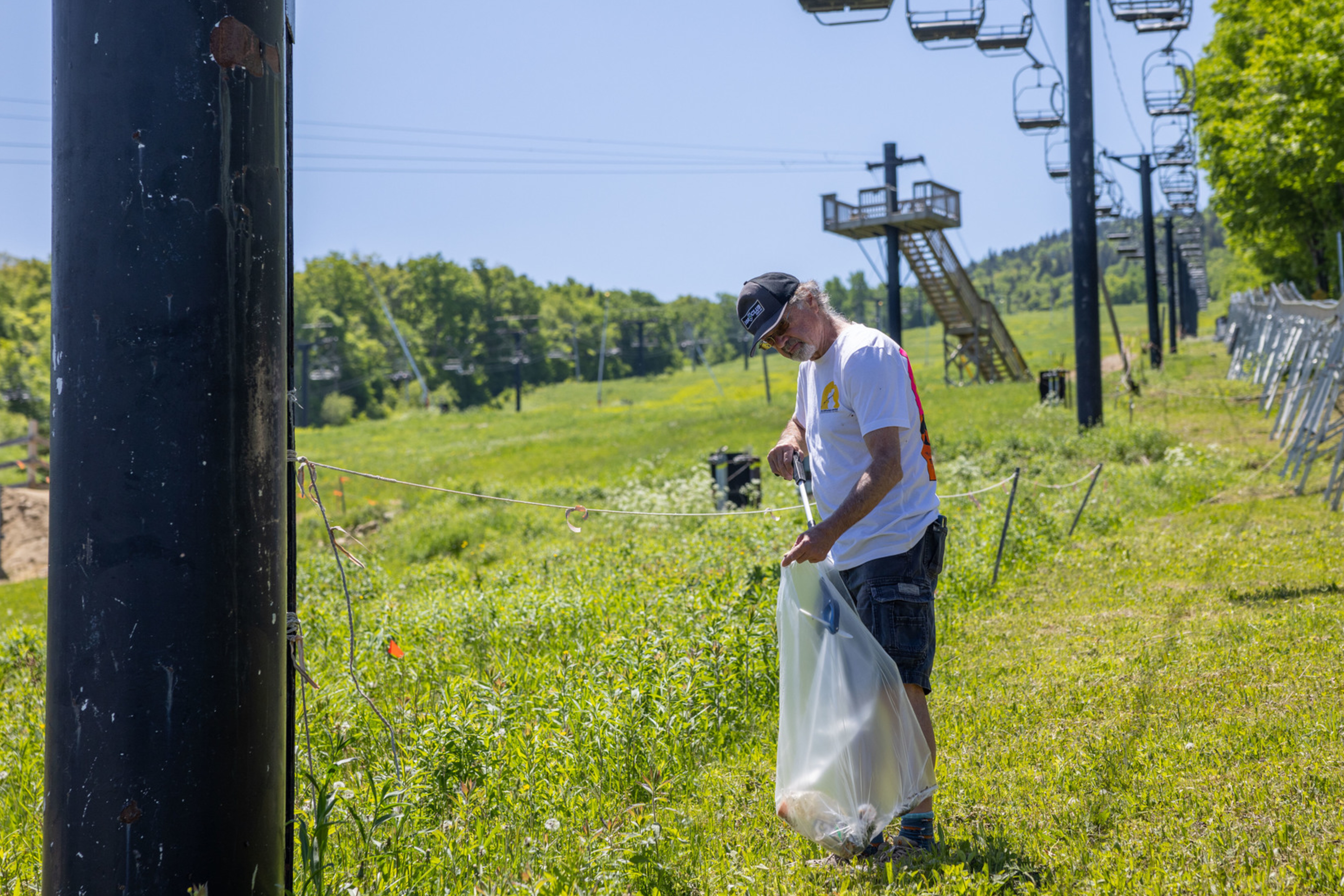 A man collects litter in a field near ski lift equipment on a sunny day.