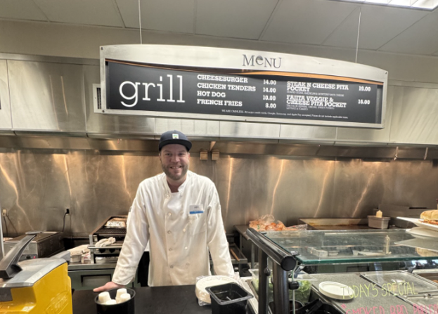 Man in a white chef coat stands in front of a grill menu featuring various foods.
