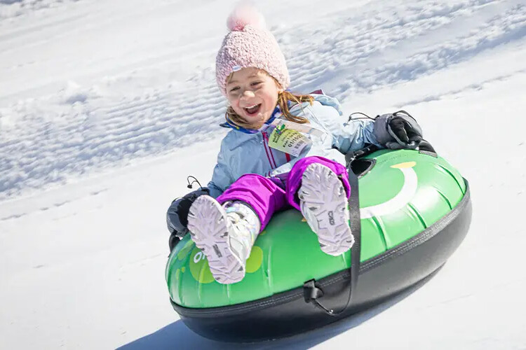 Girl joyfully sledding down a snowy hill on a green snow tube, wearing winter attire.