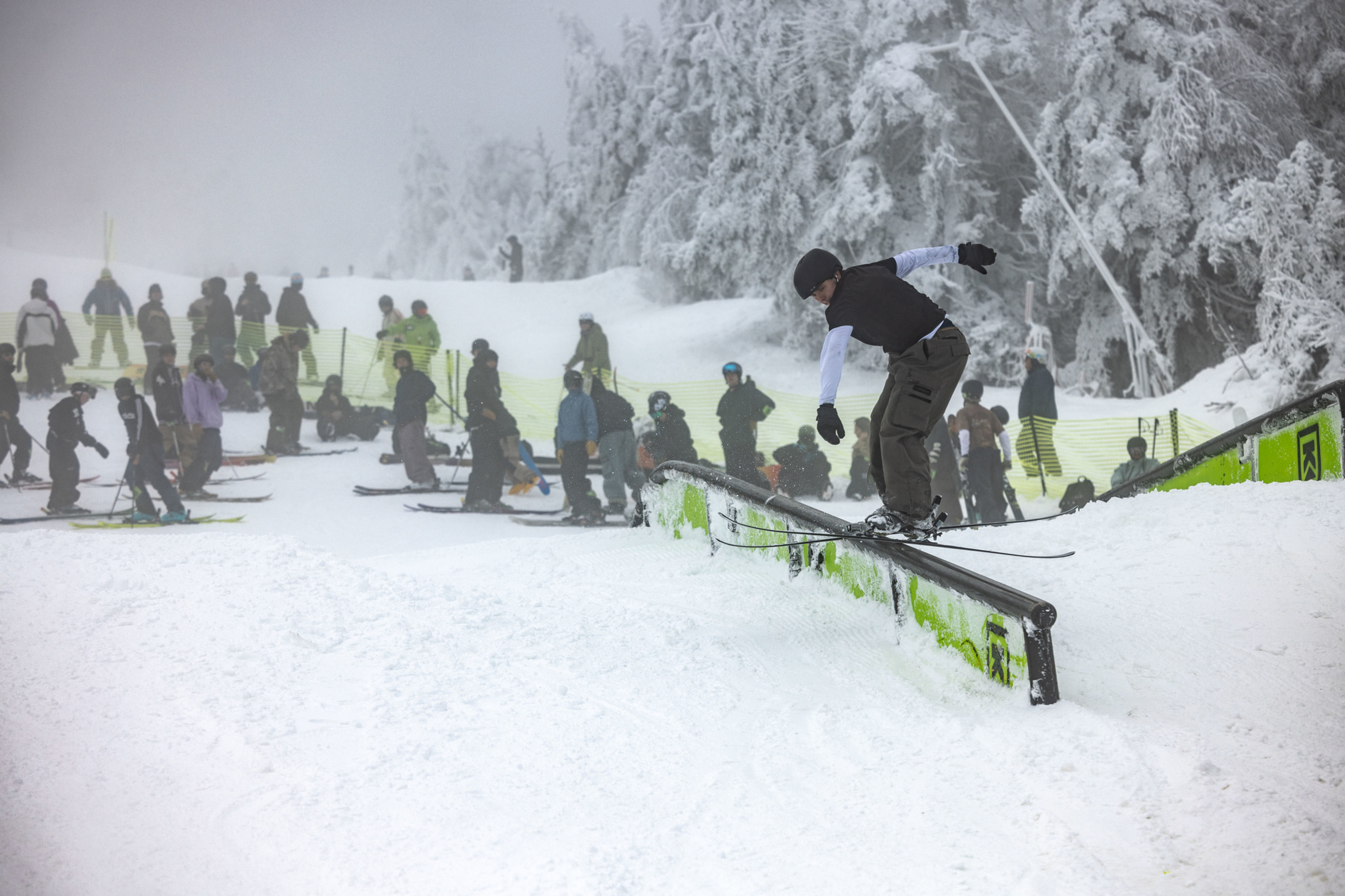 A skier performs tricks on a rail amidst a crowd on a snowy slope.
