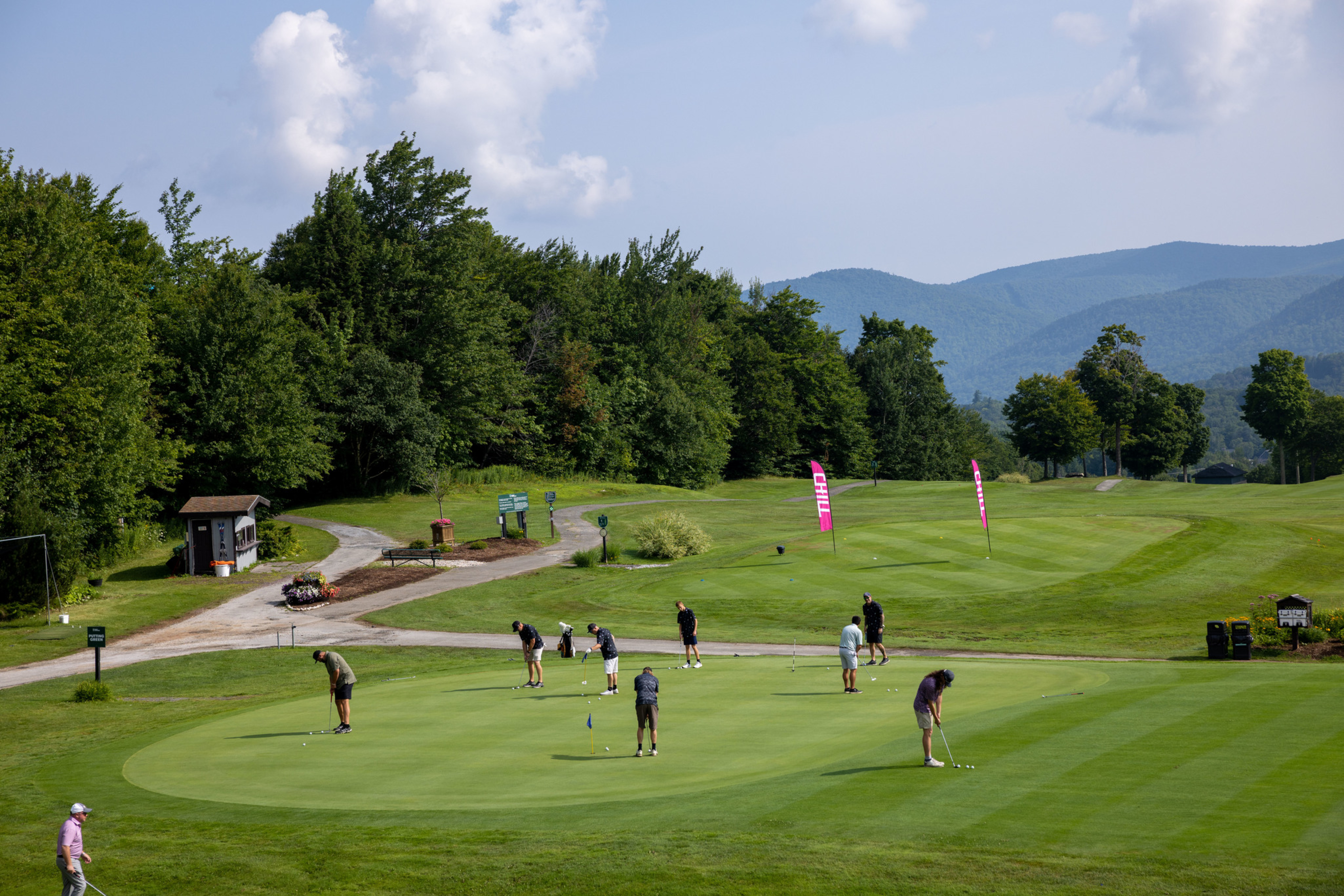 Golfers putting on a green with mountains and trees in the background.