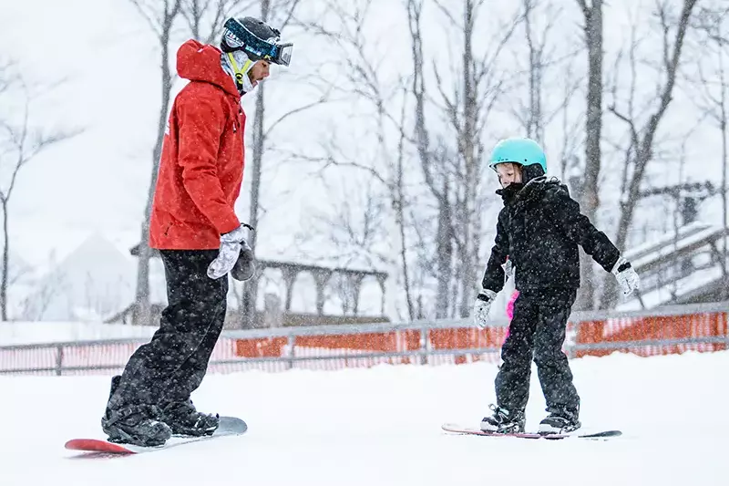 A ski instructor teaches a child to snowboard in a snowy landscape.