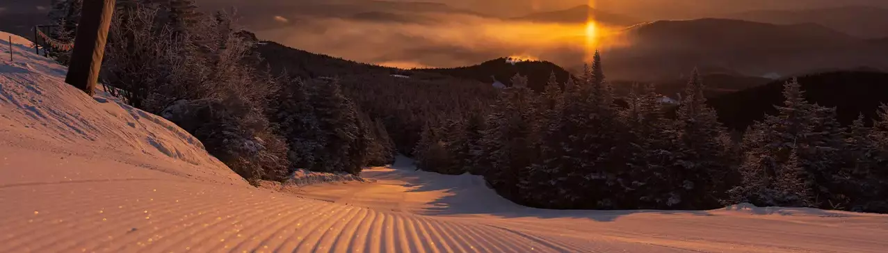 Sunset over a snowy slope, surrounded by trees and misty mountains in the distance.