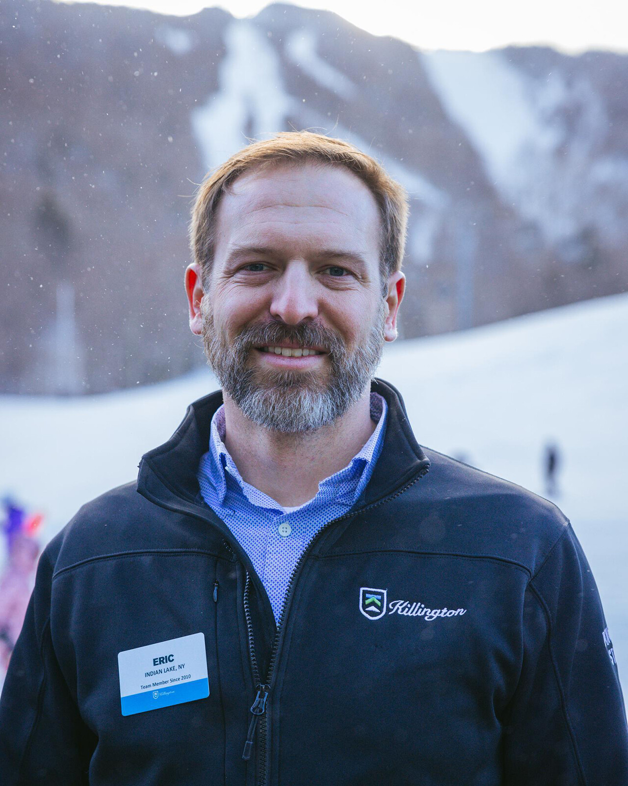 Man with beard in a black jacket stands in snowy mountain landscape.