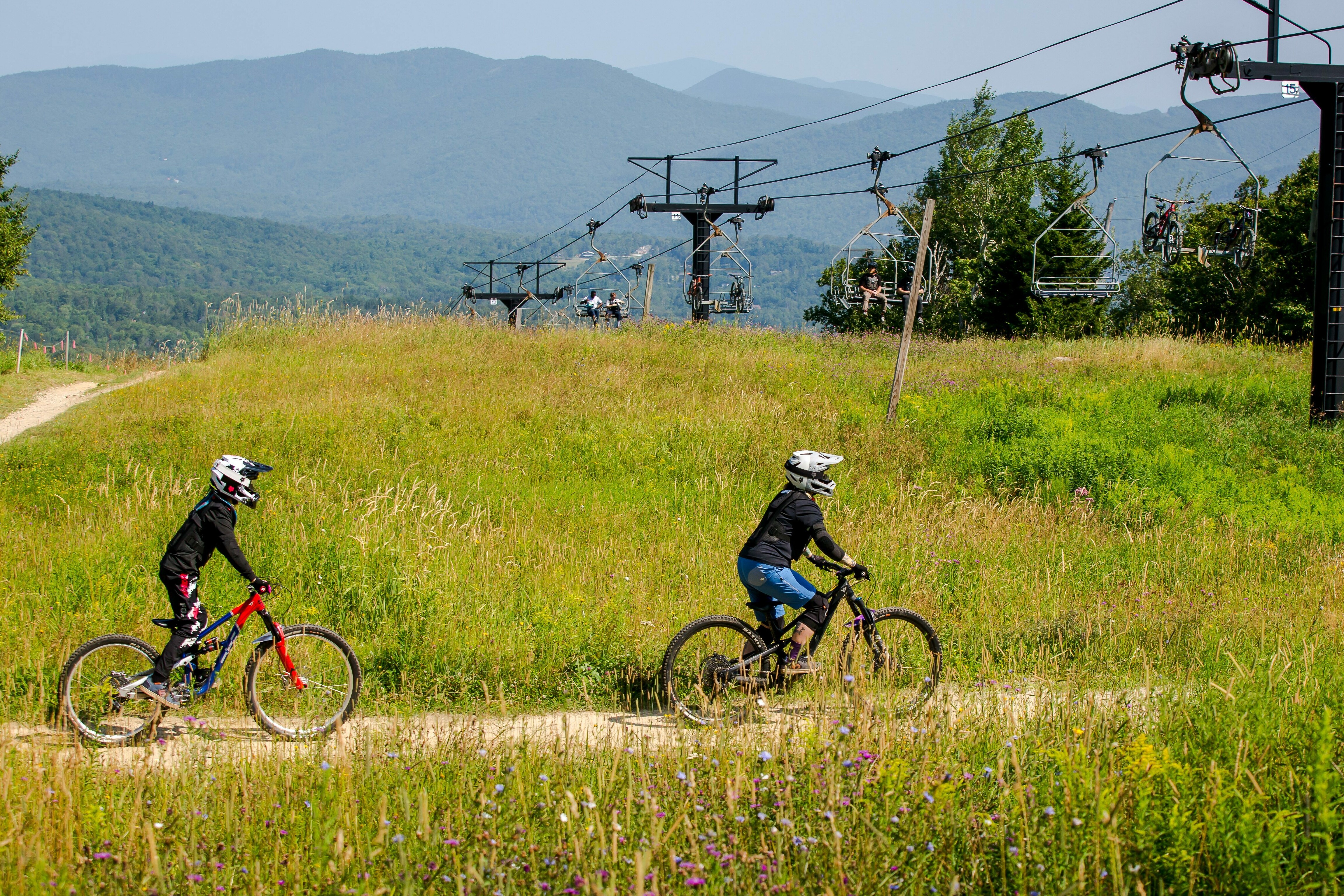 Two people ride mountain bikes on a grassy trail with a ski lift in the background.