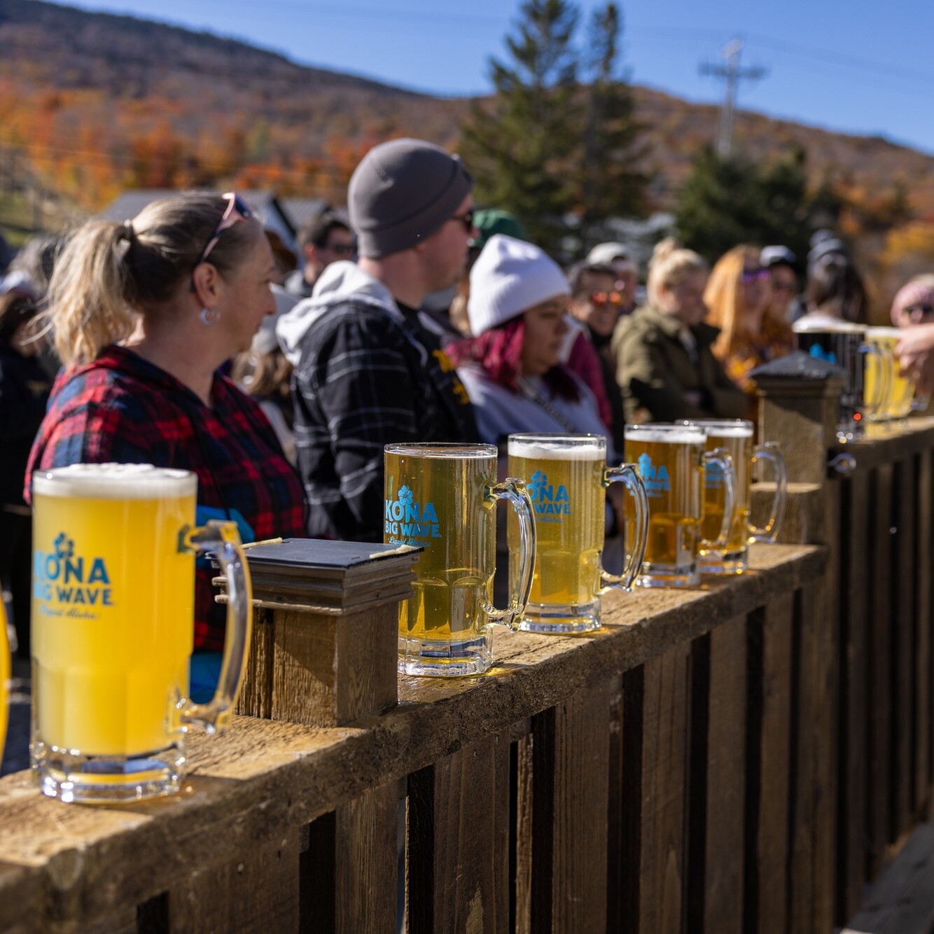 A row of beers on a wooden bar with a crowd enjoying a sunny outdoor event at Killington Mountain.