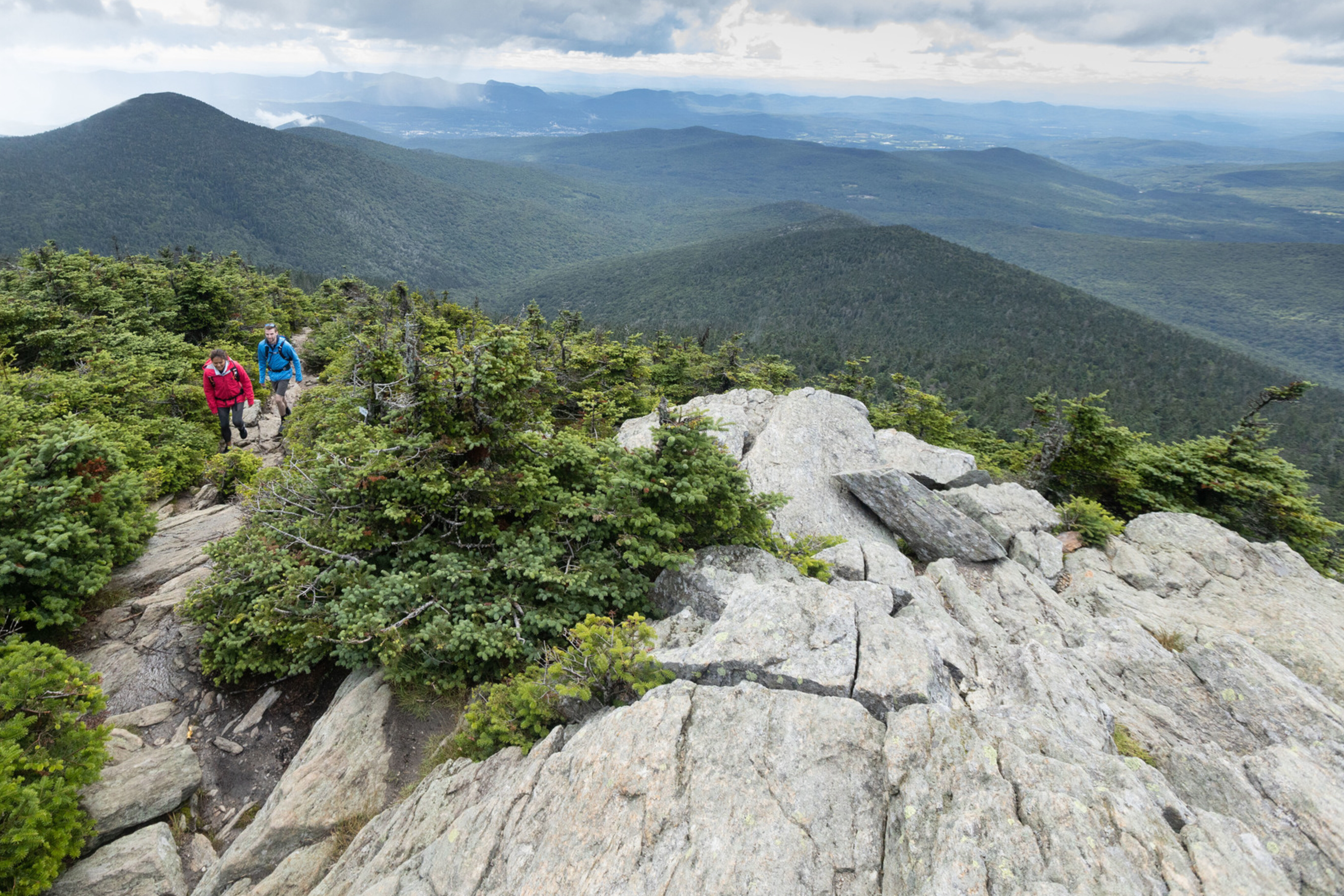 Two hikers ascend rocky terrain with lush green hills and mountains in the background.