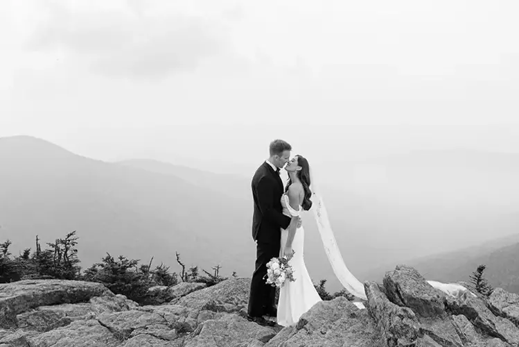 A couple kisses on a rocky mountain top, surrounded by misty mountains.