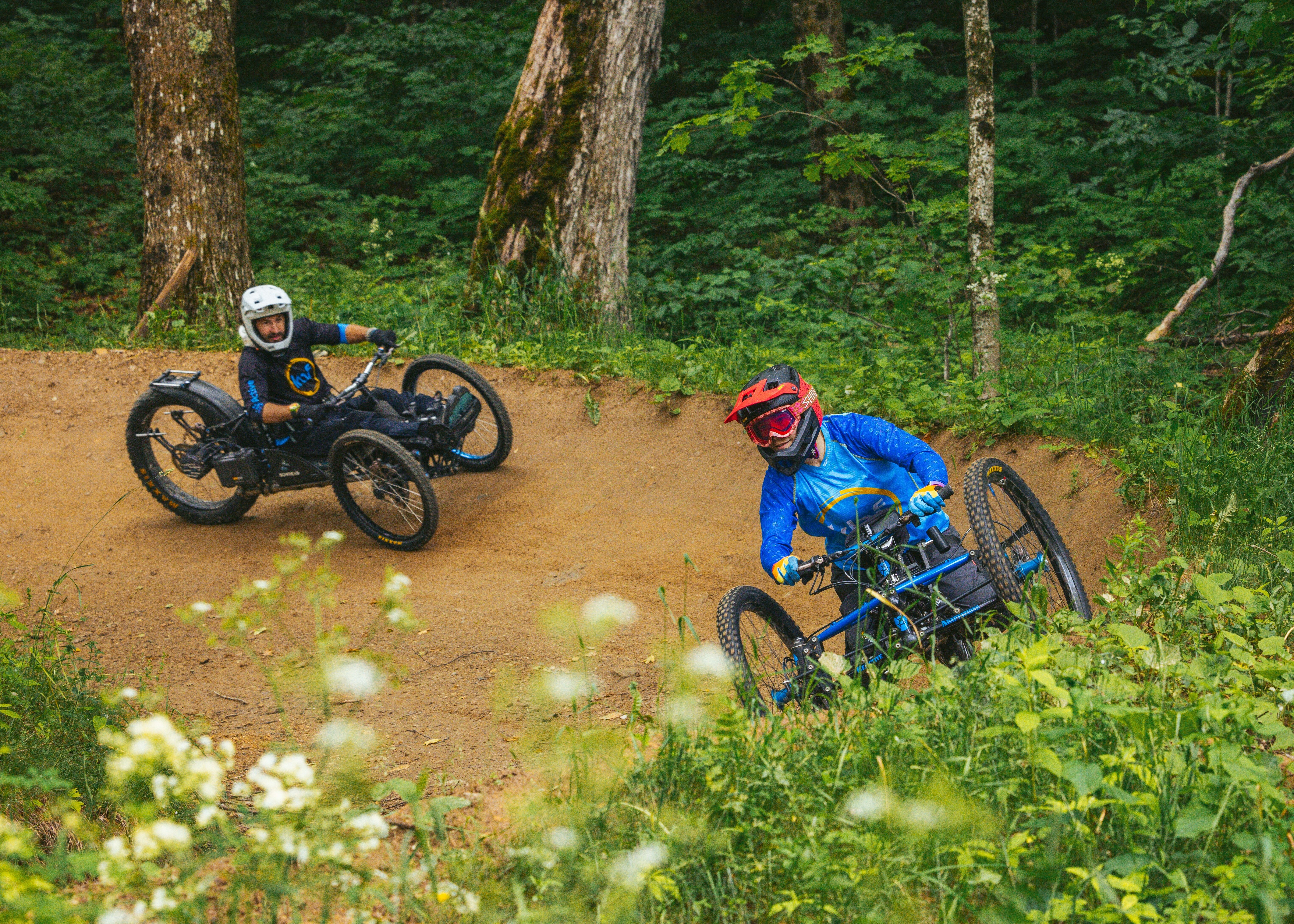 Two riders navigate a dirt turn on adaptive mountain bikes in a forested area.