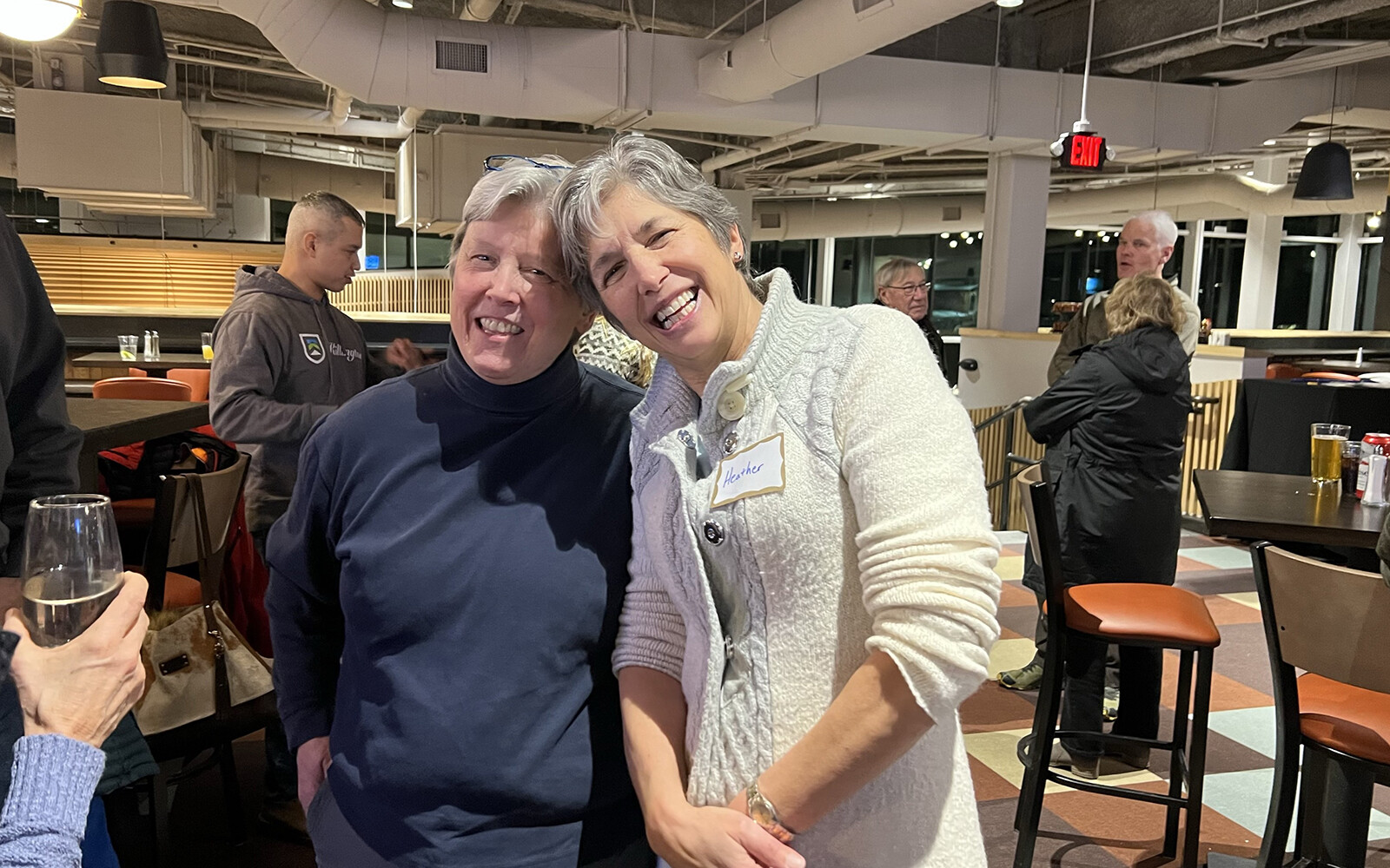 Two smiling women pose together in a lively indoor setting.