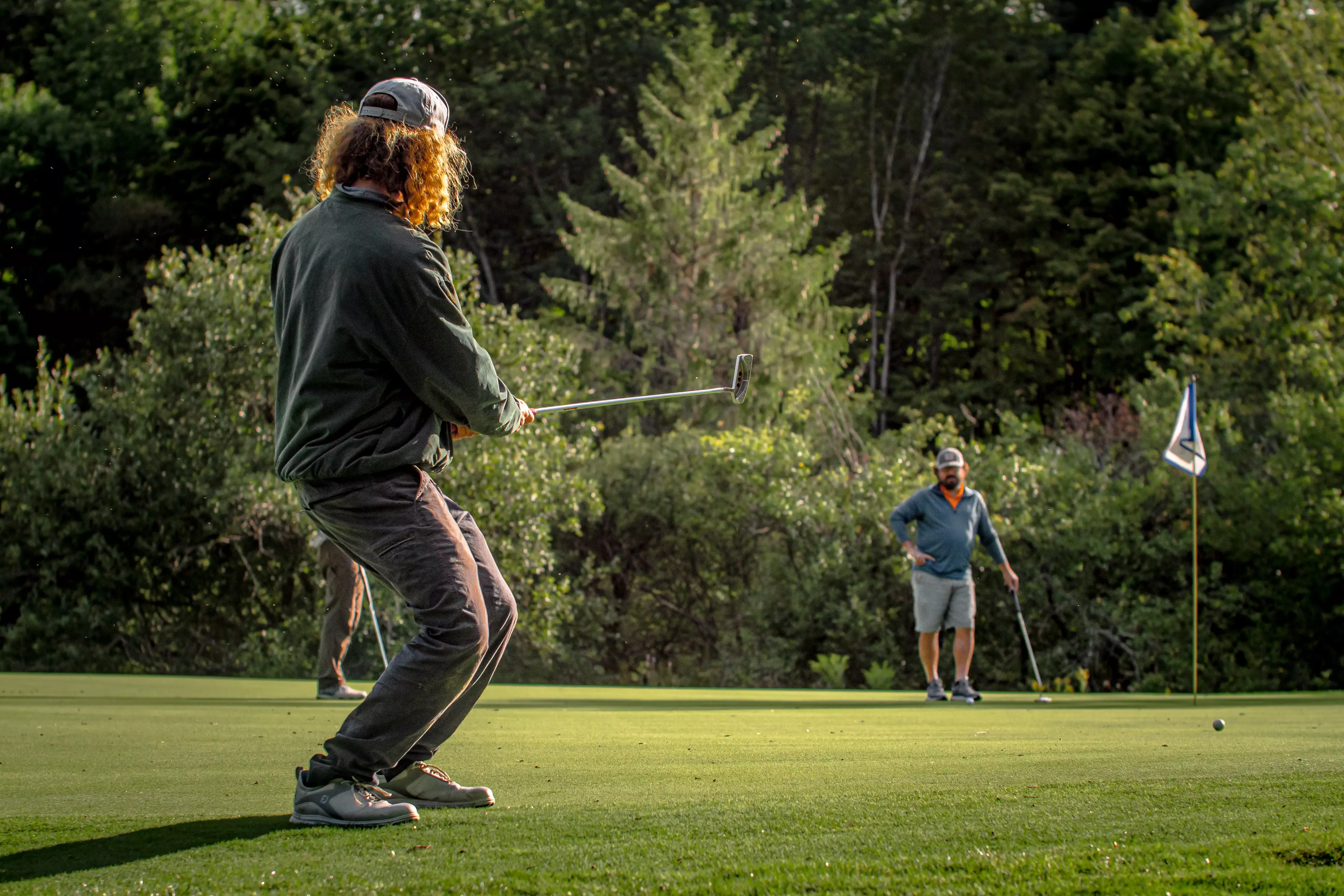Two golfers on a green, one preparing to putt while the other observes.