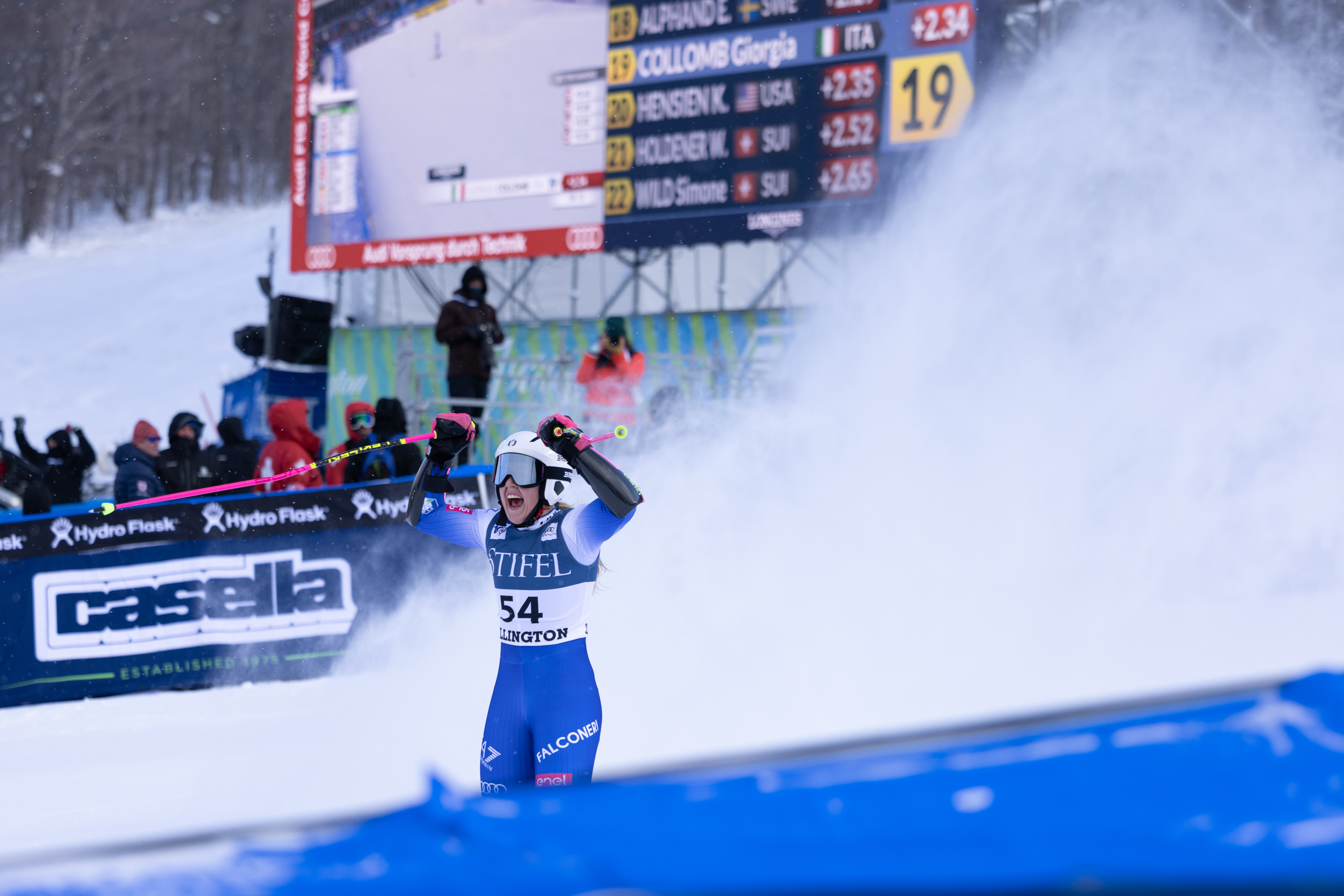 A skier celebrates with raised poles amid snow, with a scoreboard in the background.