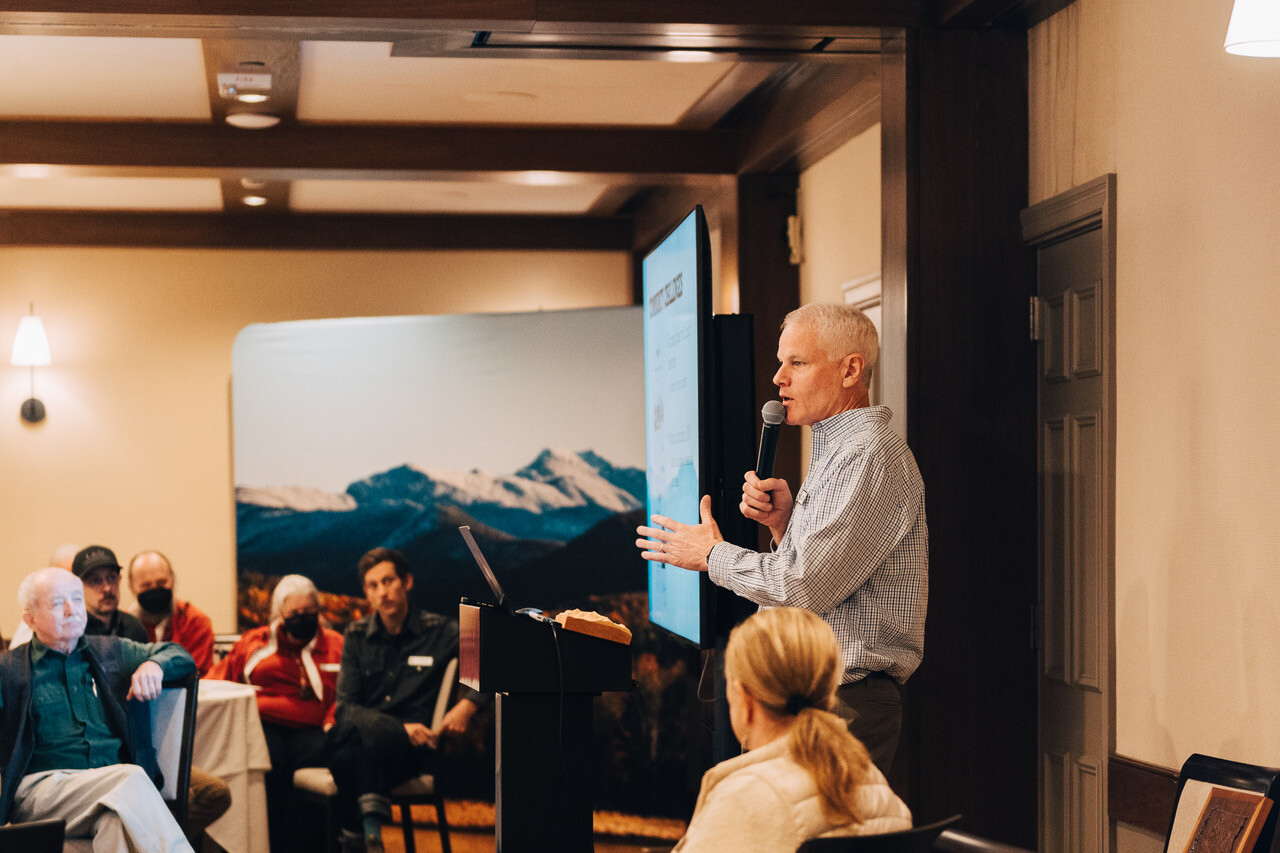 A speaker addresses an audience in a conference room, with a mountain backdrop.