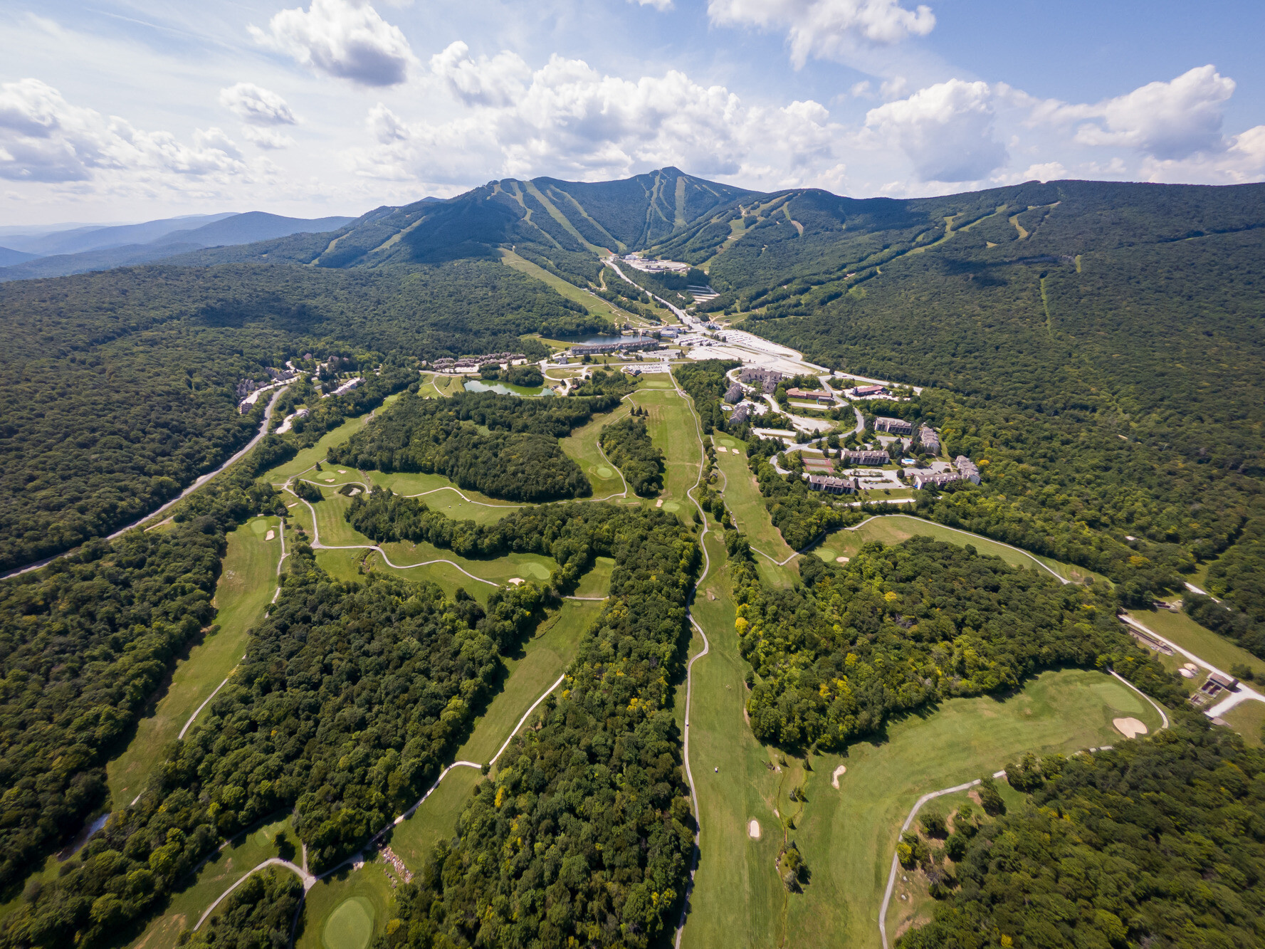 Aerial view of lush green hills, golf courses, and ski slopes under a blue sky.