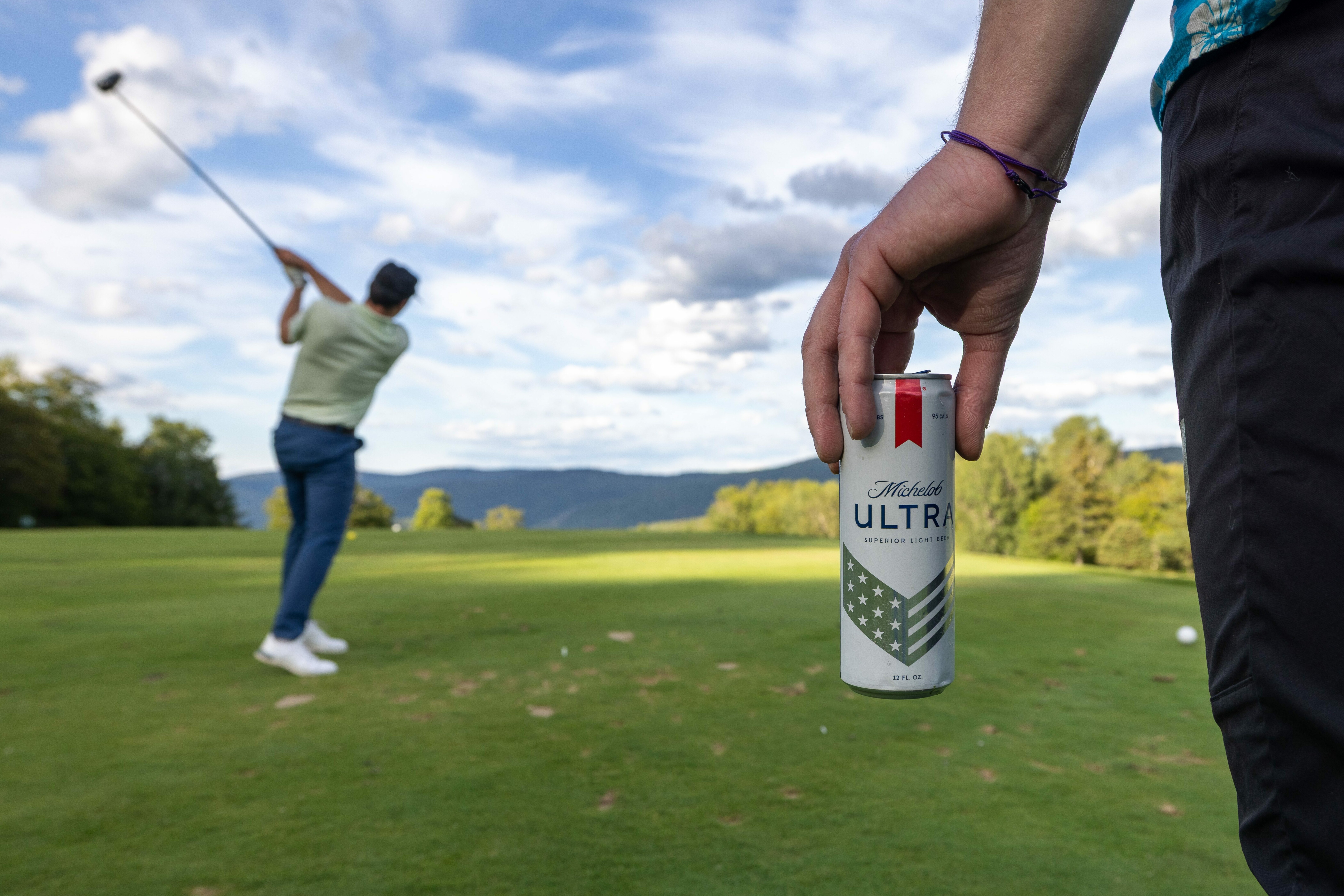 A golfer swings while someone holds a Michelob ULTRA beer can nearby.