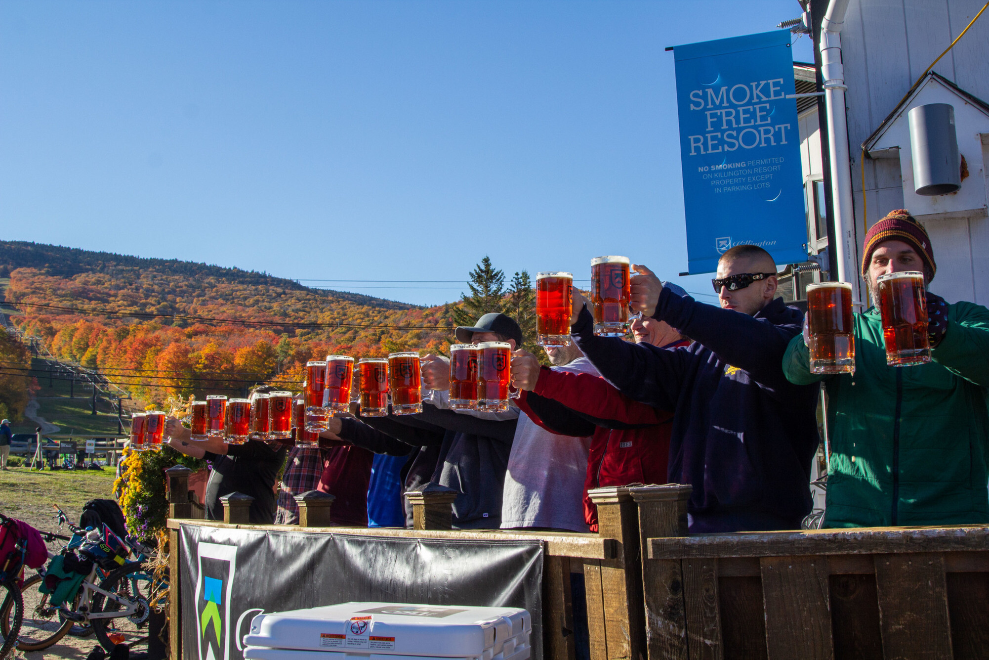 A group of people raising mugs of beer outdoors with autumn foliage in the background.