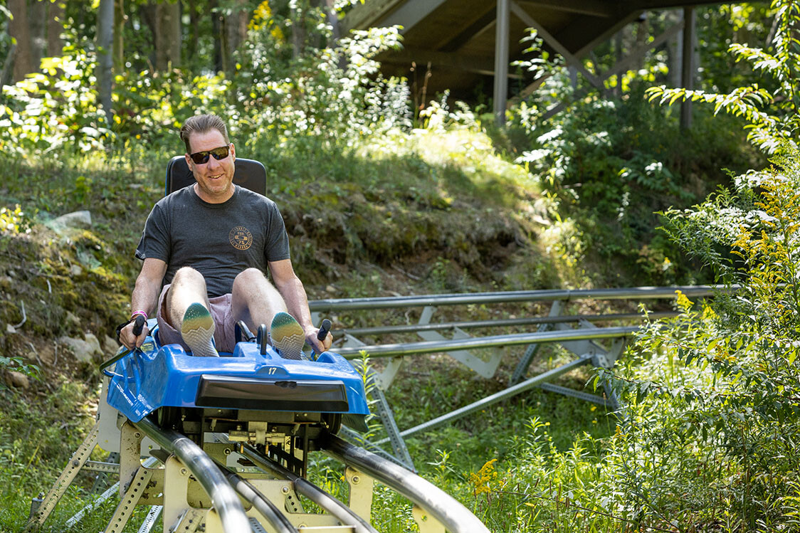 Person riding a blue alpine coaster track through a wooded area, smiling and relaxed.
