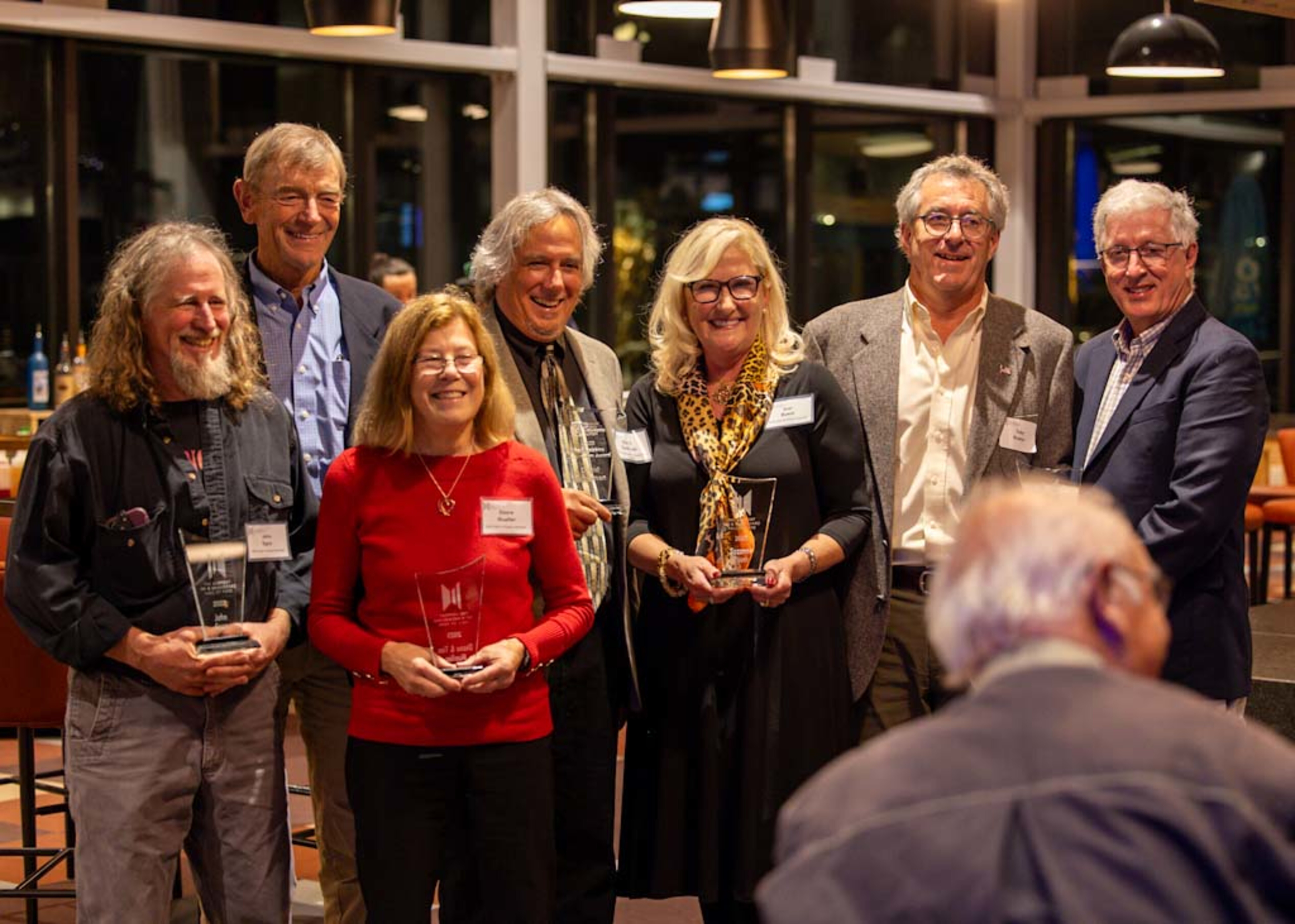 Group of seven award recipients posing together at an event, holding their trophies.