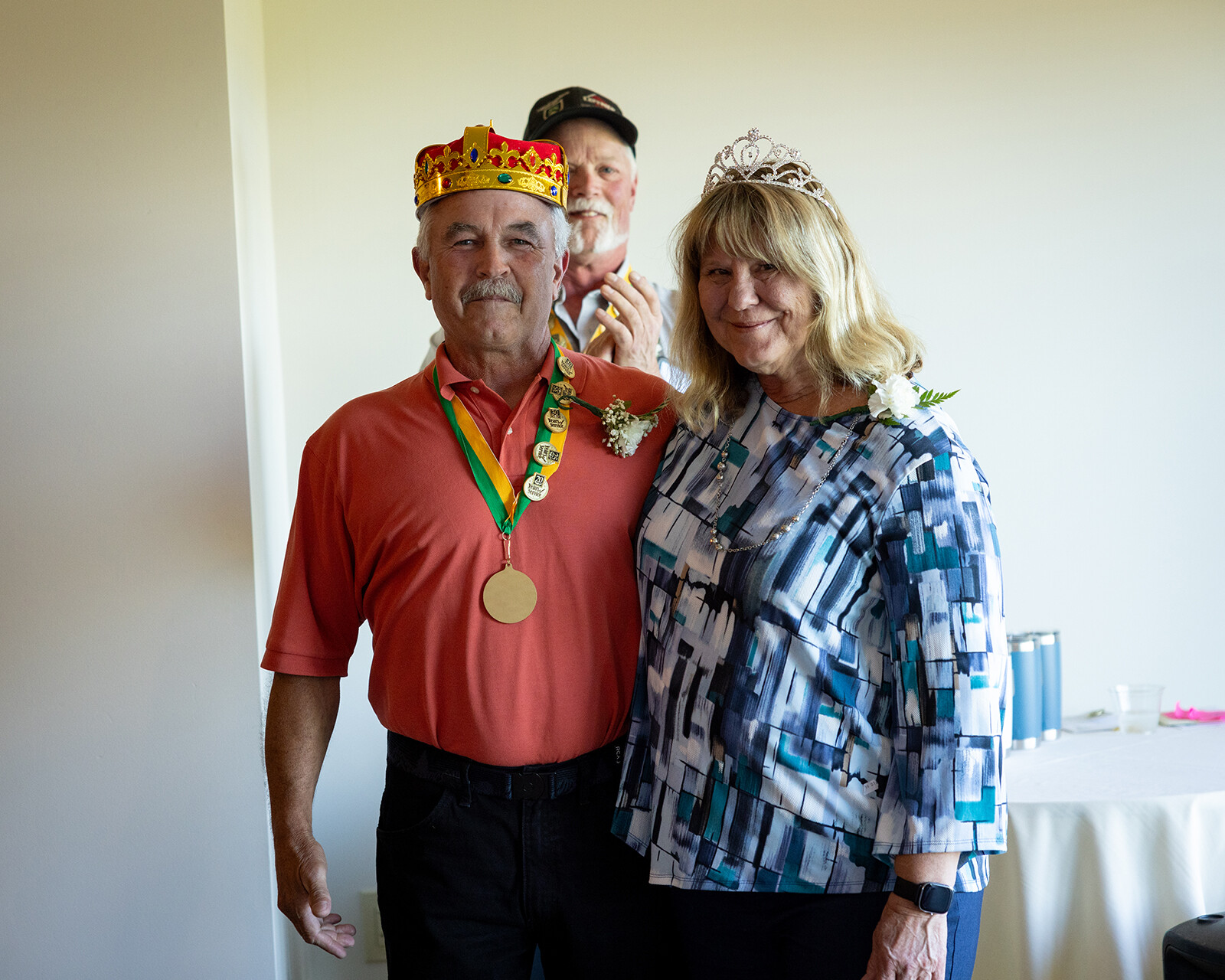 Two people proudly pose with crowns and medals; a third person smiles in the background.