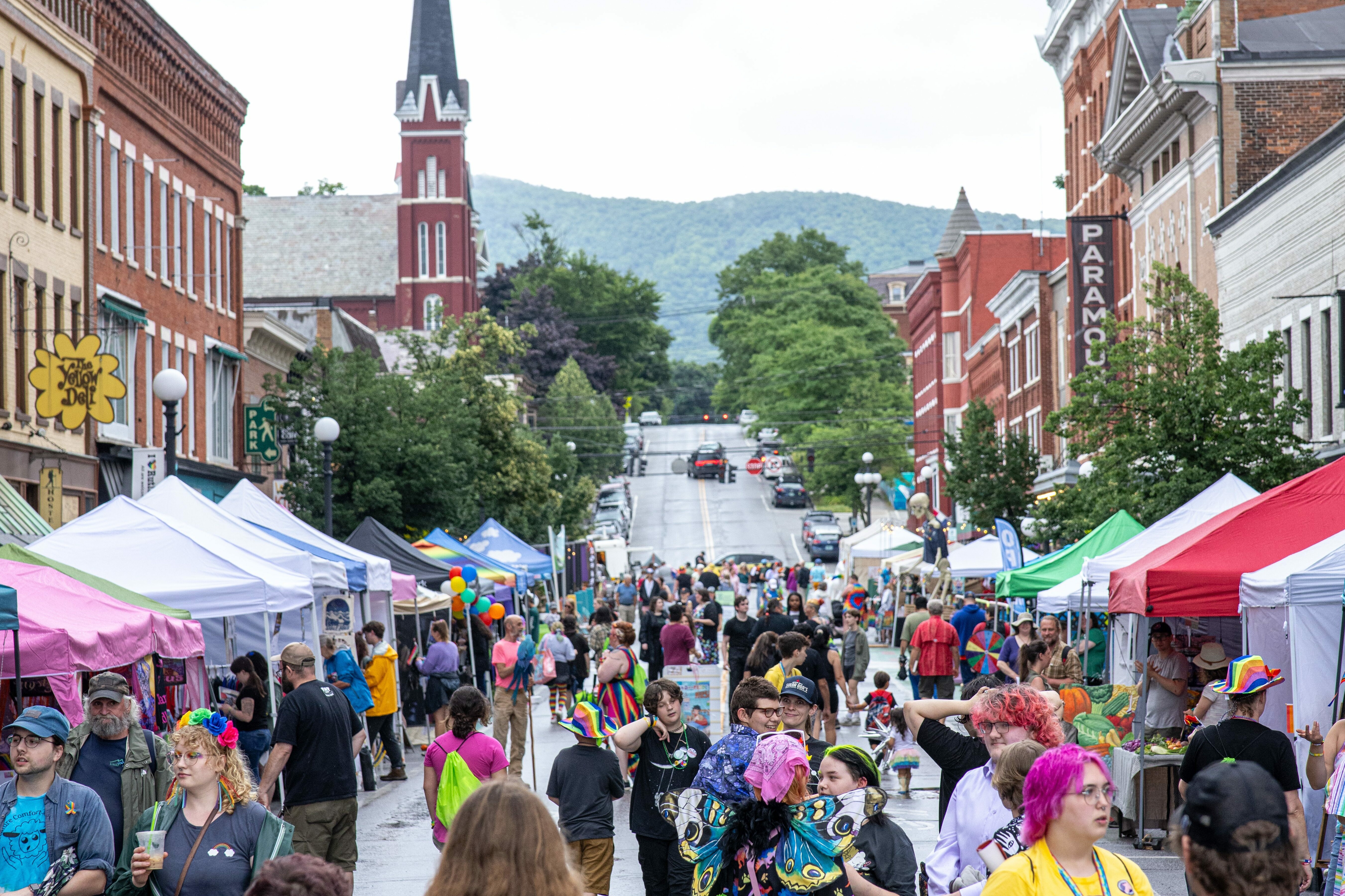 Busy street festival with colorful tents and diverse crowds, set against a mountainous backdrop.