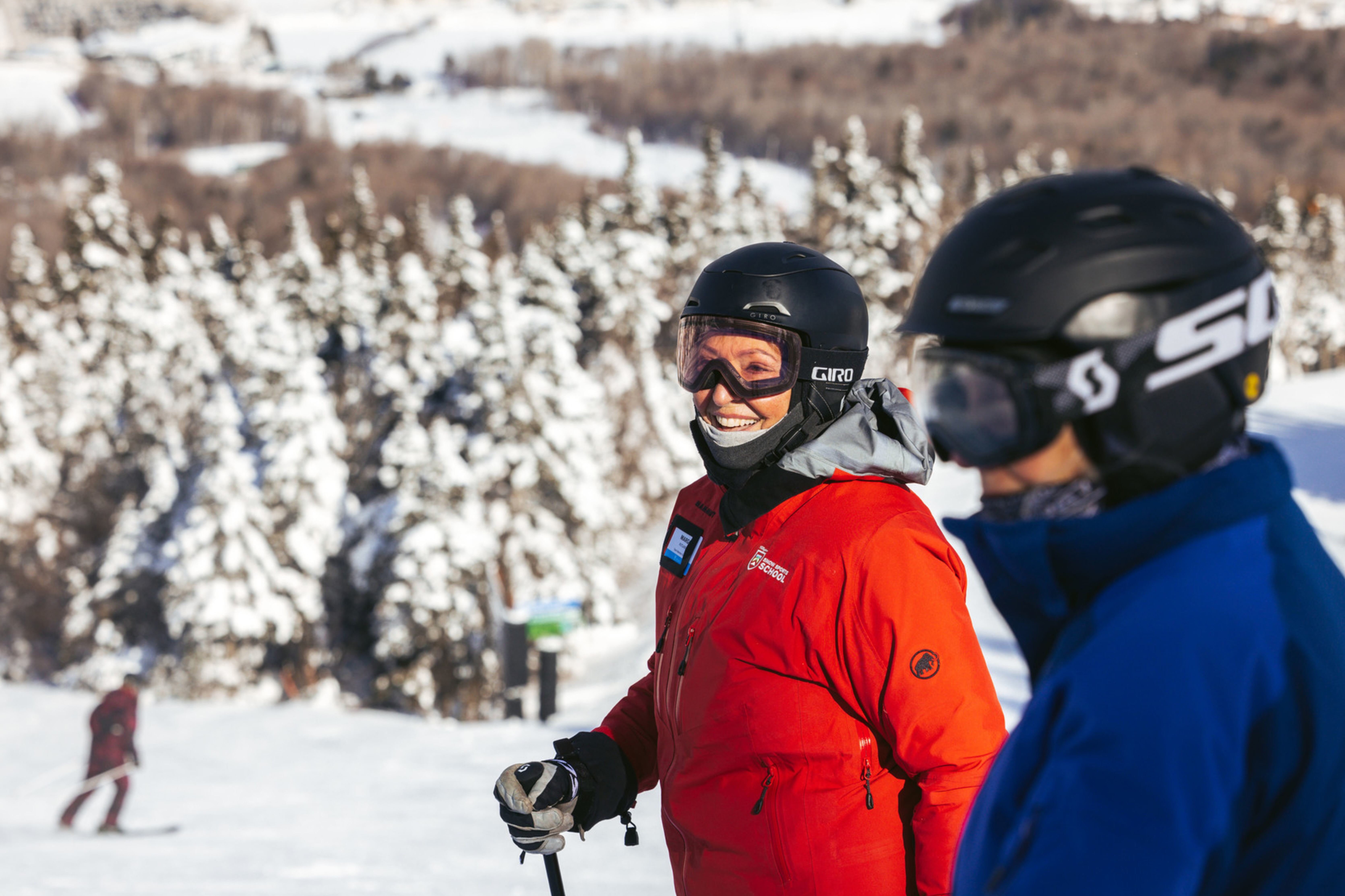 Two smiling skiers in helmets stand on a snowy slope with trees in the background.